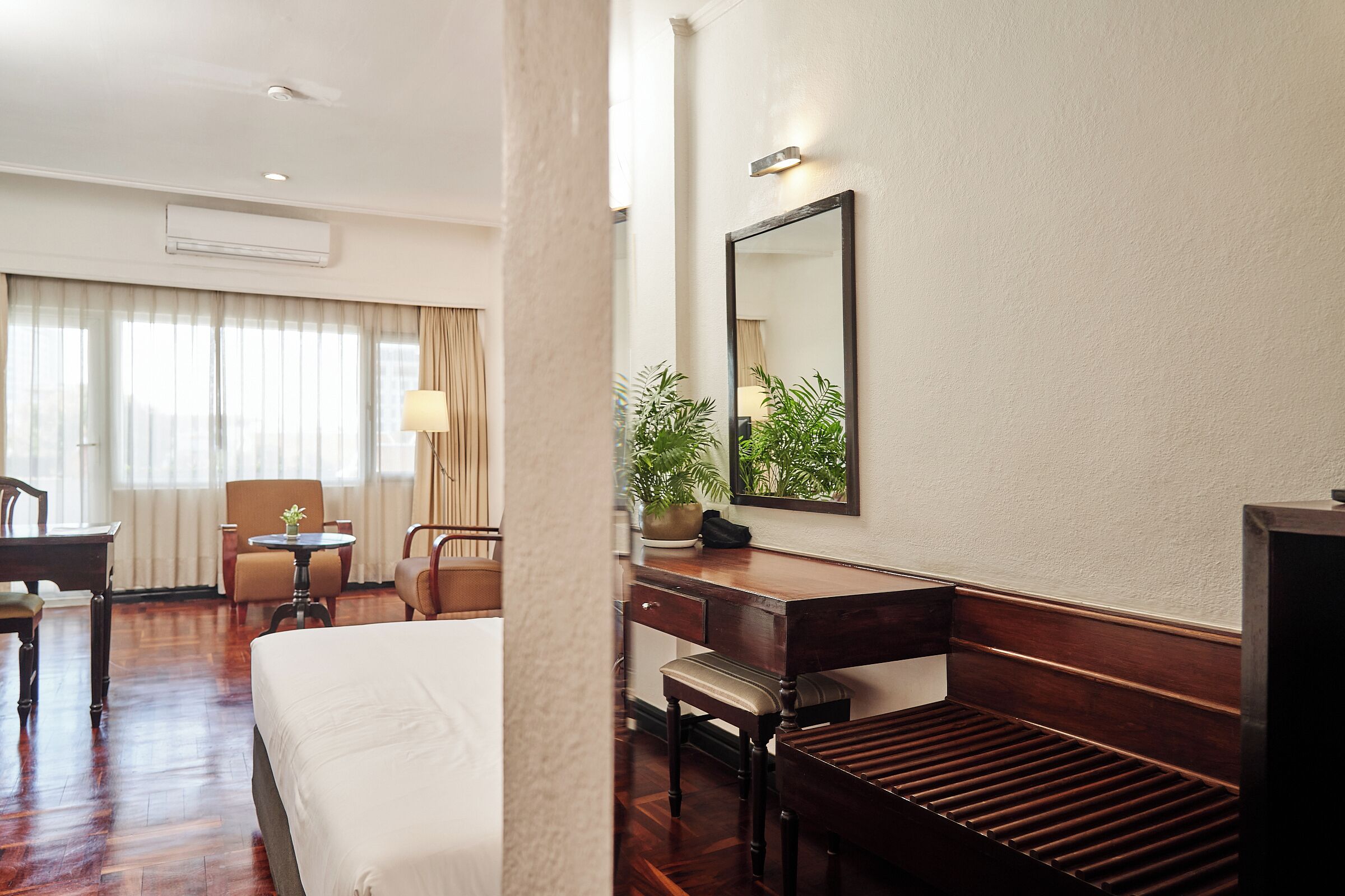 A hotel room with a bed, a desk, a chair, a small table, and a potted plant.