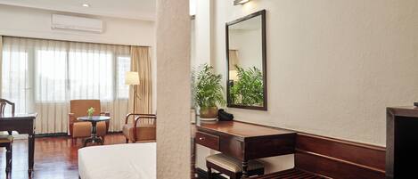 A hotel room with a bed, a desk, a chair, a small table, and a potted plant.