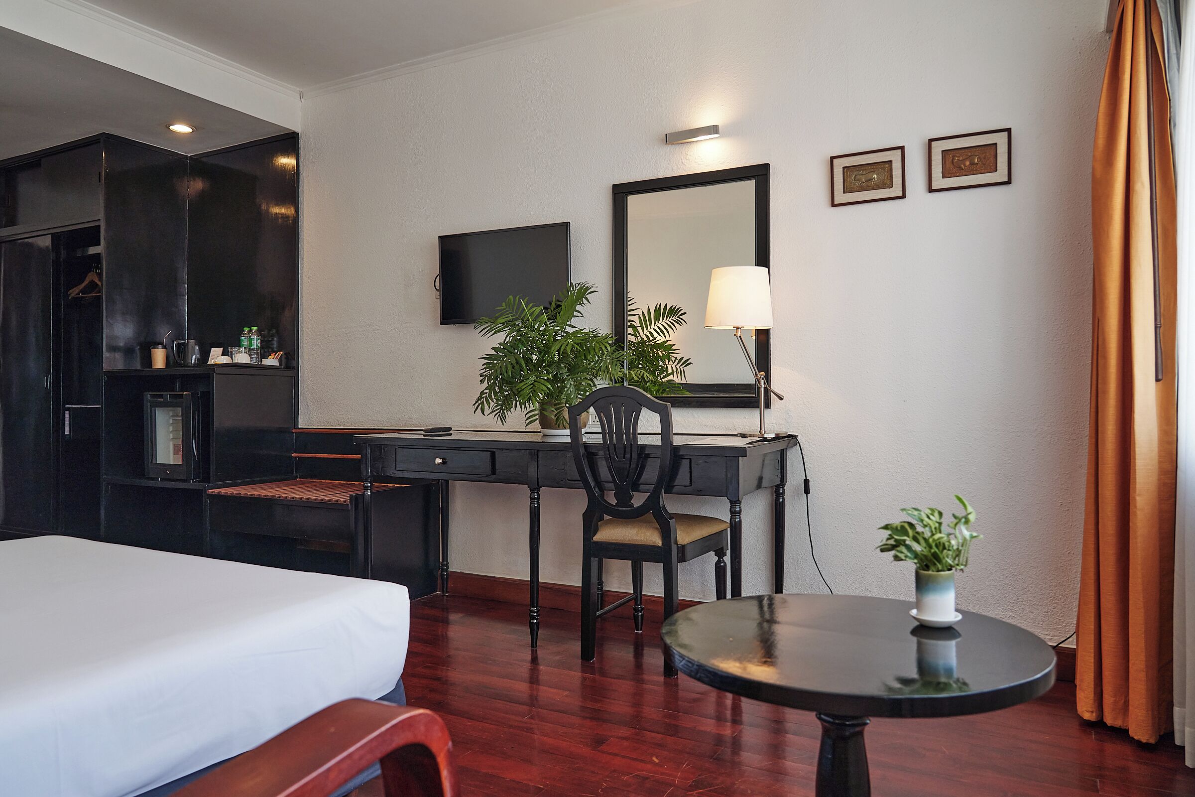 A hotel room with a bed, a desk with a chair, a television, a mirror, and a small table with a plant.