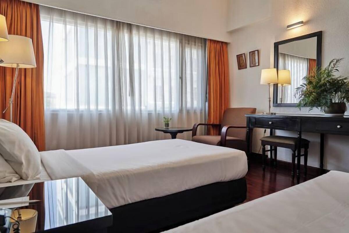Hotelkamer met twee bedden, een bureau, een stoel, een lamp, een plant en een groot raam met gordijnen.