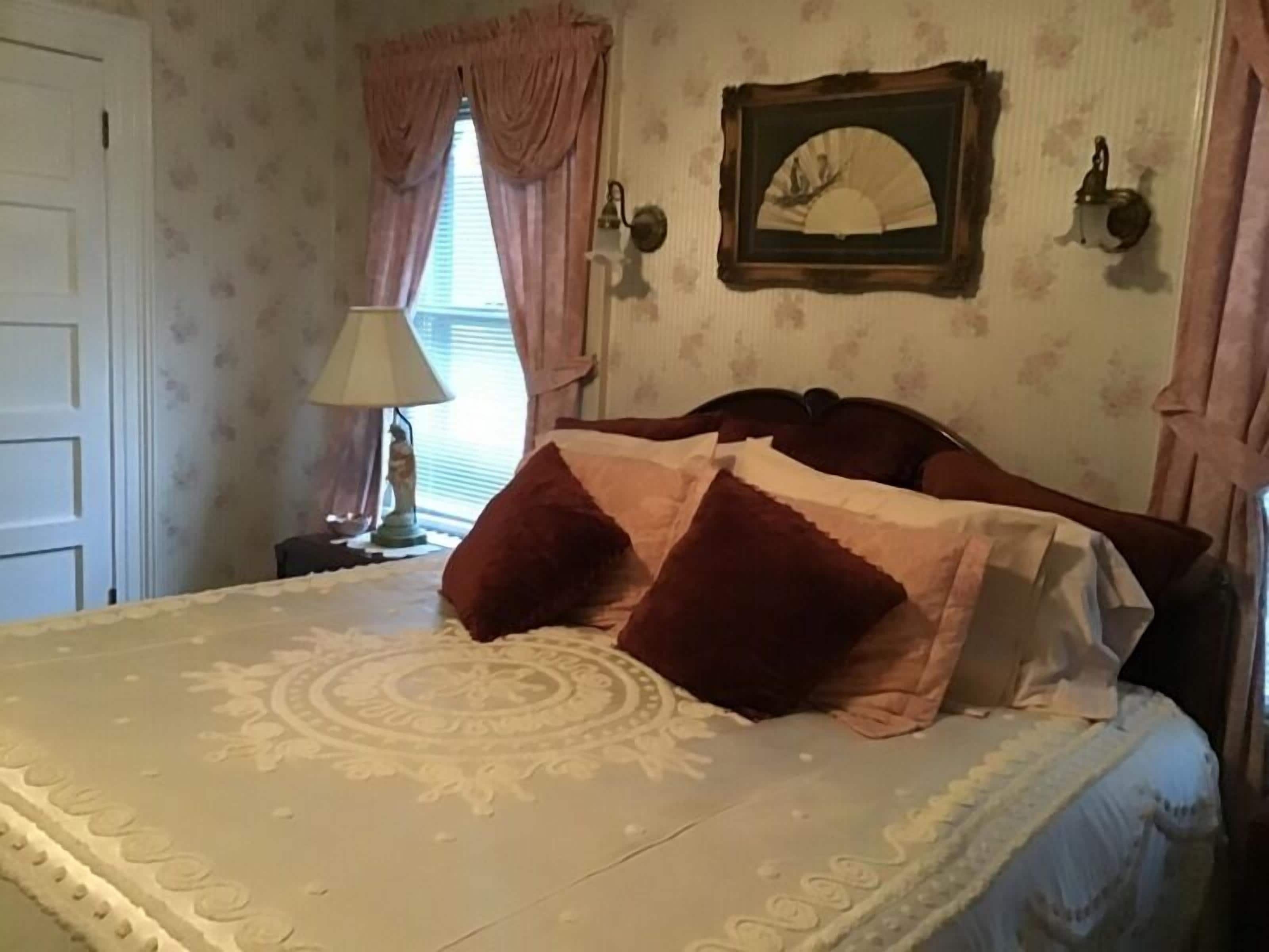 Un dormitorio con cama, mesita de noche, lámpara, ventana con cortinas y espejo adosado a la pared.