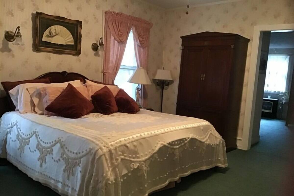 Ein Schlafzimmer mit Bett, Nachttisch, Lampe, Kleiderschrank und einem Fenster mit Vorhängen.