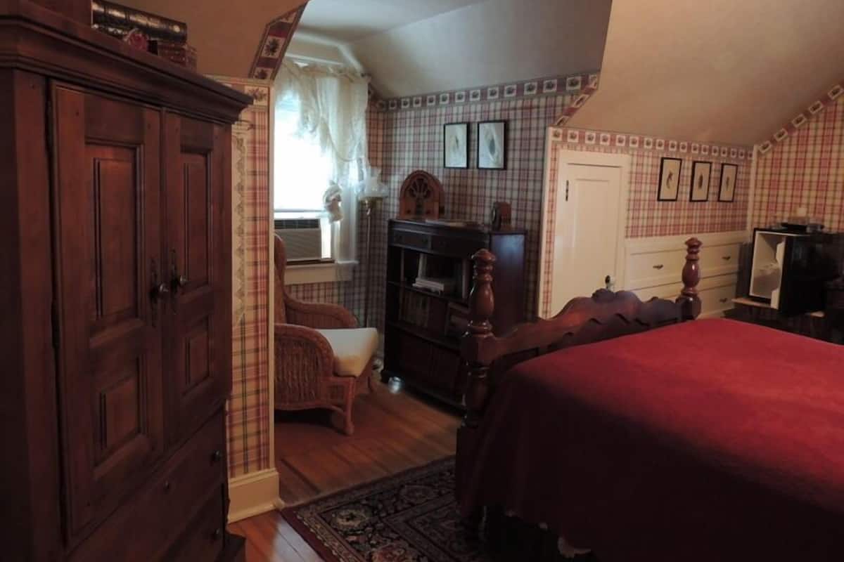 Ein Schlafzimmer mit einem Holzschrank, einem Bett mit roter Decke, einem Sessel, einer Kommode und einem Spiegel.