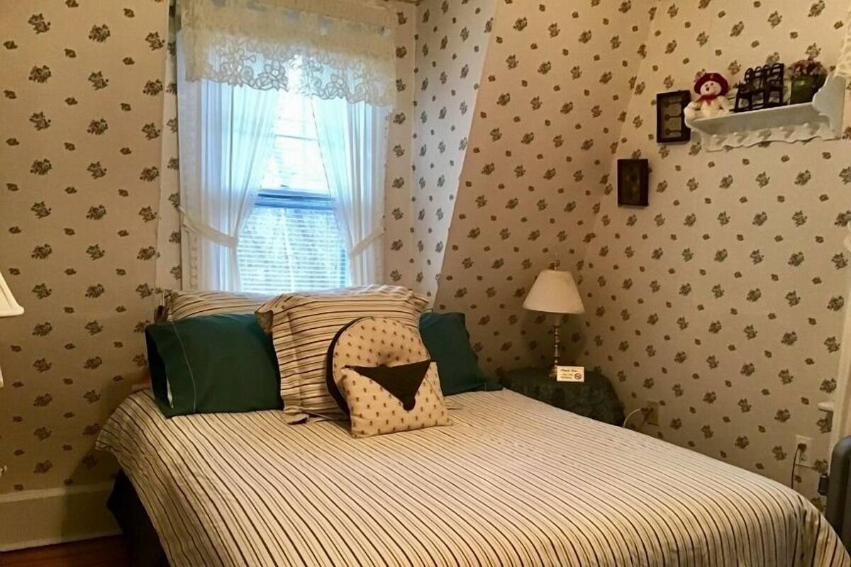 Ein Schlafzimmer mit gestreifter Bettdecke, grünen Kissen, einem Spitzenfenster und floralem Wandpapier.