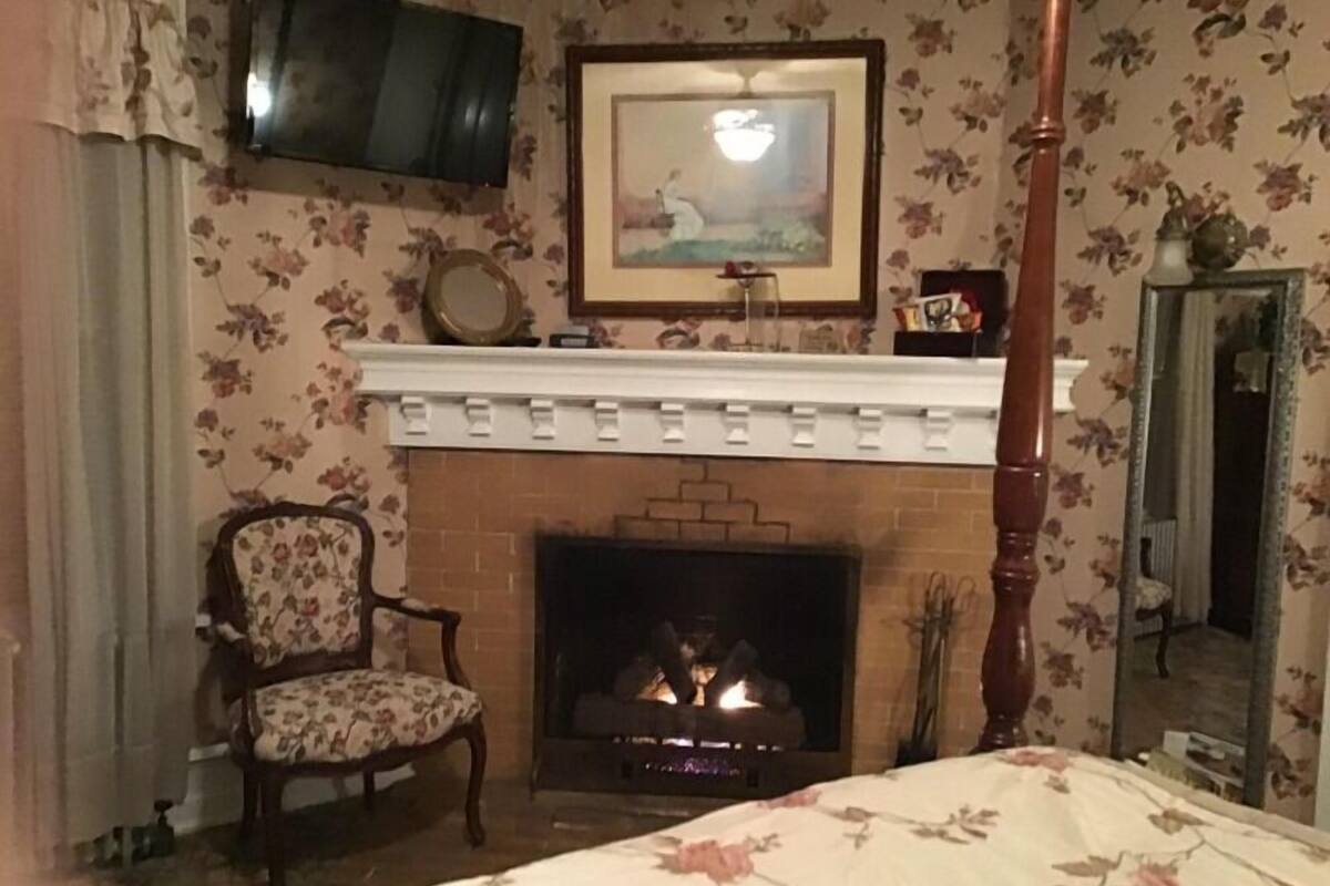 Ein Schlafzimmer mit Kamin, Sessel, Bett und Fernseher, an den Wänden blumengemustertes Papier.