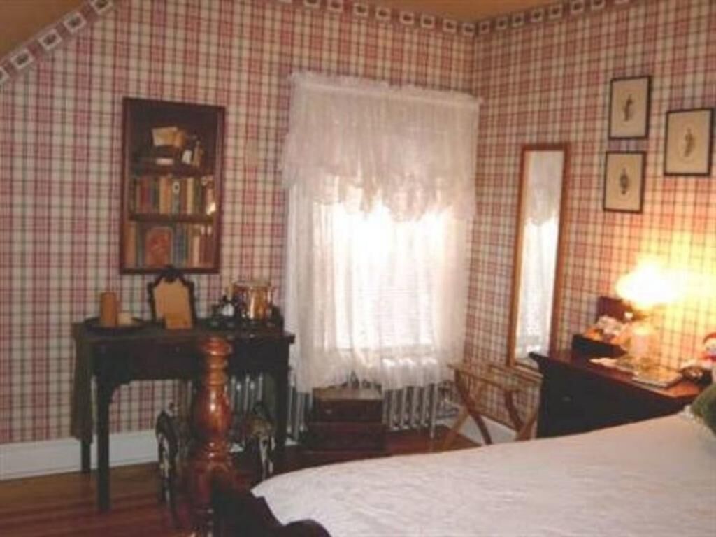 A room with a checkered wallpaper, a bed with a white bedspread, a wooden chair, a small table with a lamp, and a bookshelf.