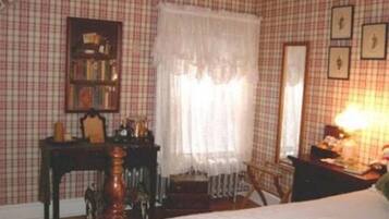 A room with a checkered wallpaper, a bed with a white bedspread, a wooden chair, a small table with a lamp, and a bookshelf.