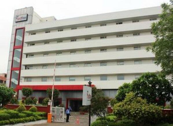 Un hotel de varios pisos con fachada roja y blanca, múltiples balcones y un área de entrada ajardinada.