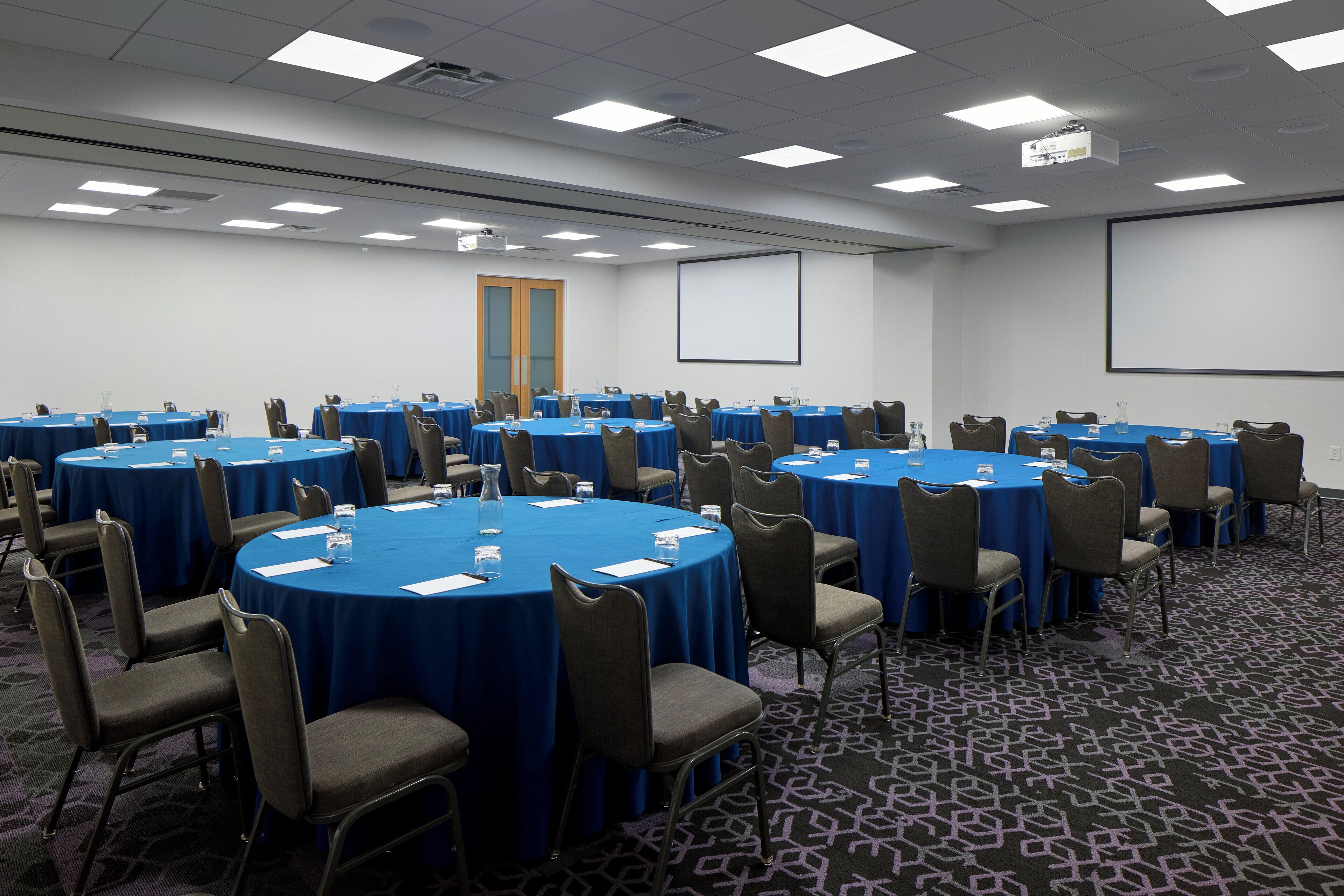 A conference room with multiple round tables covered with blue tablecloths, arranged in rows. Each table has a name card, water glass, and a notepad. The room has a carpeted floor, a projector screen, and a door in the background.