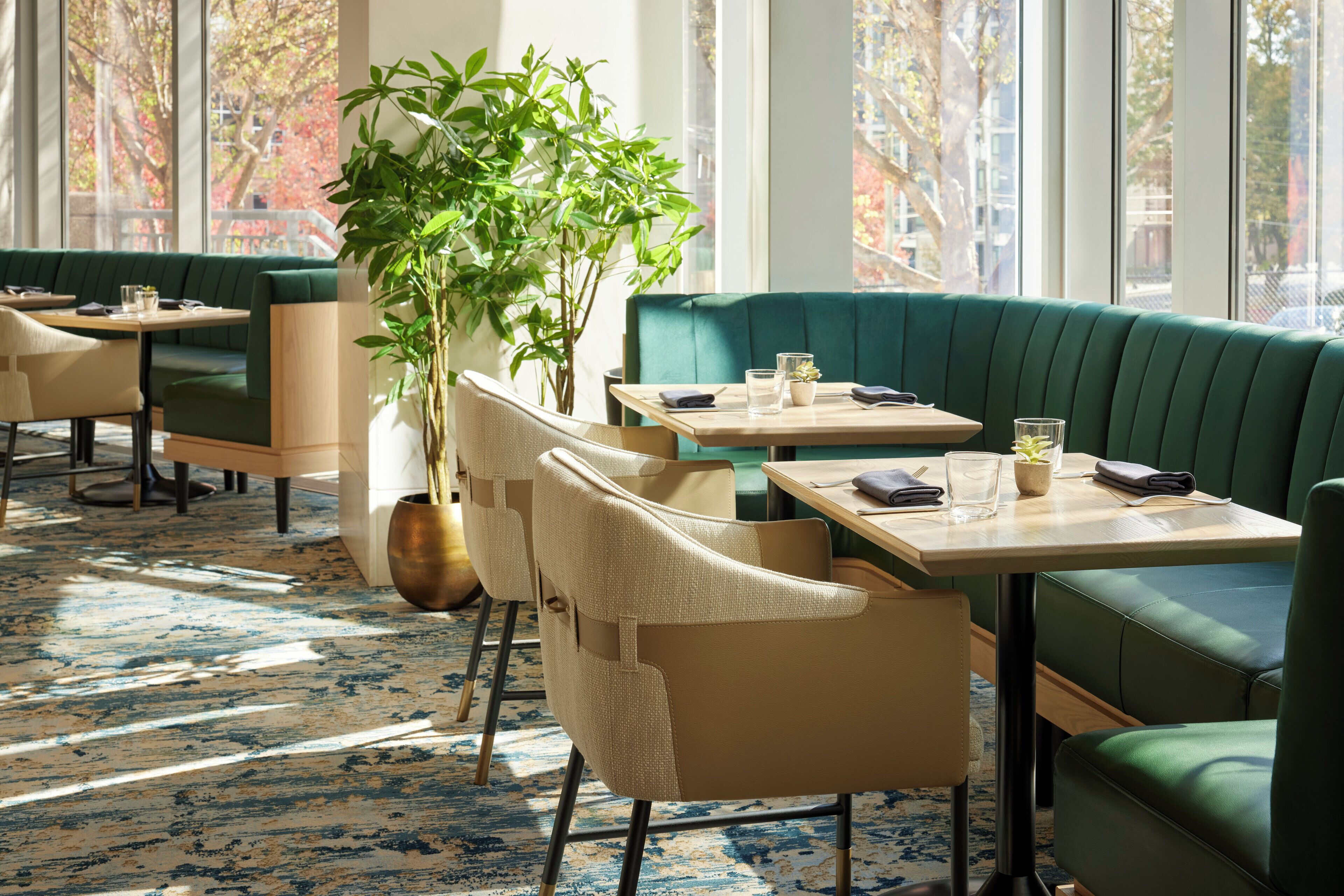 A restaurant with green booths and beige chairs, a large plant, and a patterned carpet.