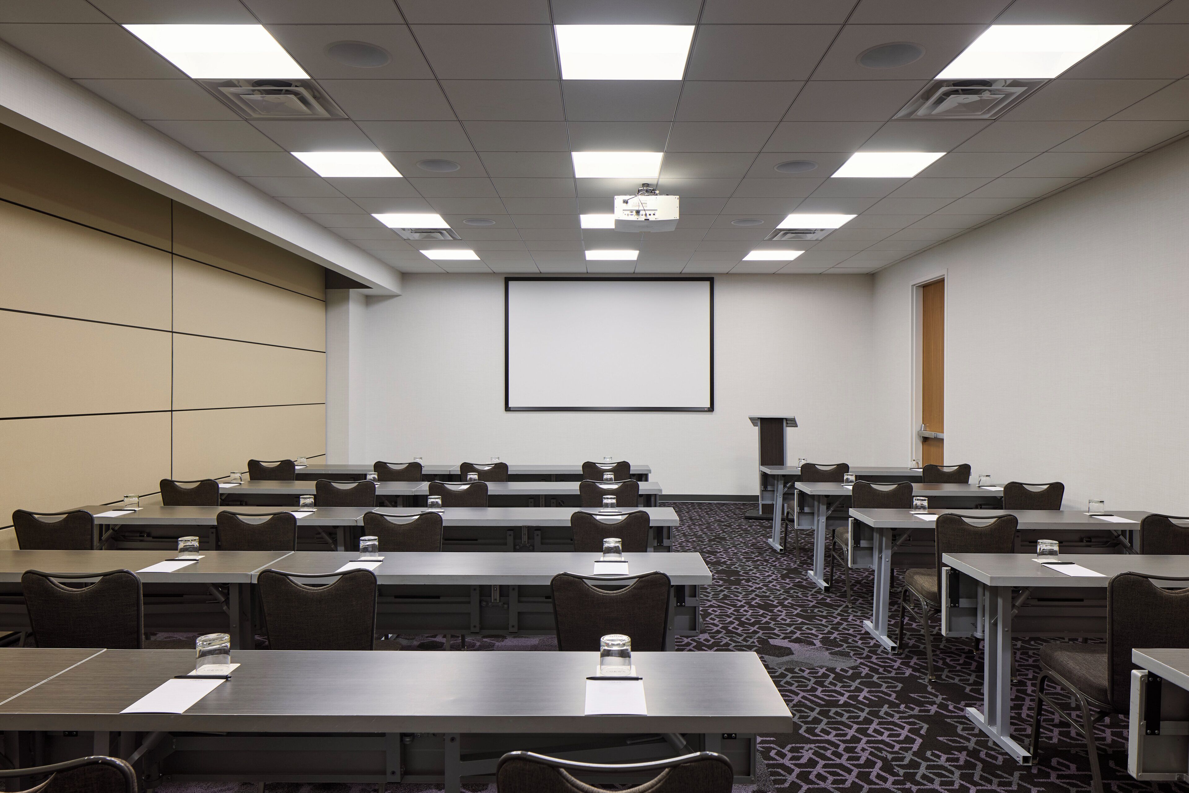 Une salle de conférence dotée de plusieurs tables et chaises, d’un écran de projection et d’un sol recouvert de moquette.