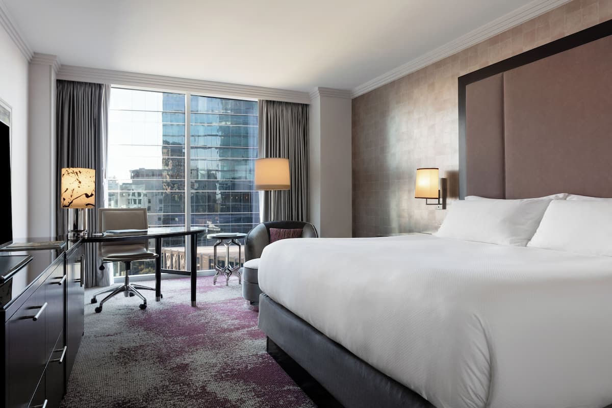 A hotel room with a large bed, a desk, a chair, and a view of a cityscape through the window.