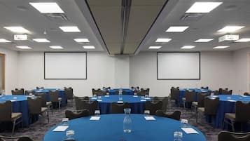 Sala de conferências com mesas redondas, cadeiras e tela de projeção.
