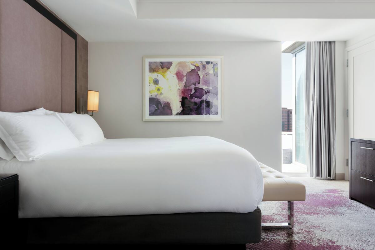 A modern hotel room with a large bed, a bedside table, a lamp, and a window with curtains.