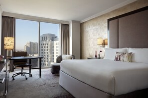 A hotel room with a large bed, a desk, and a chair. There is a window with a view of buildings outside.
