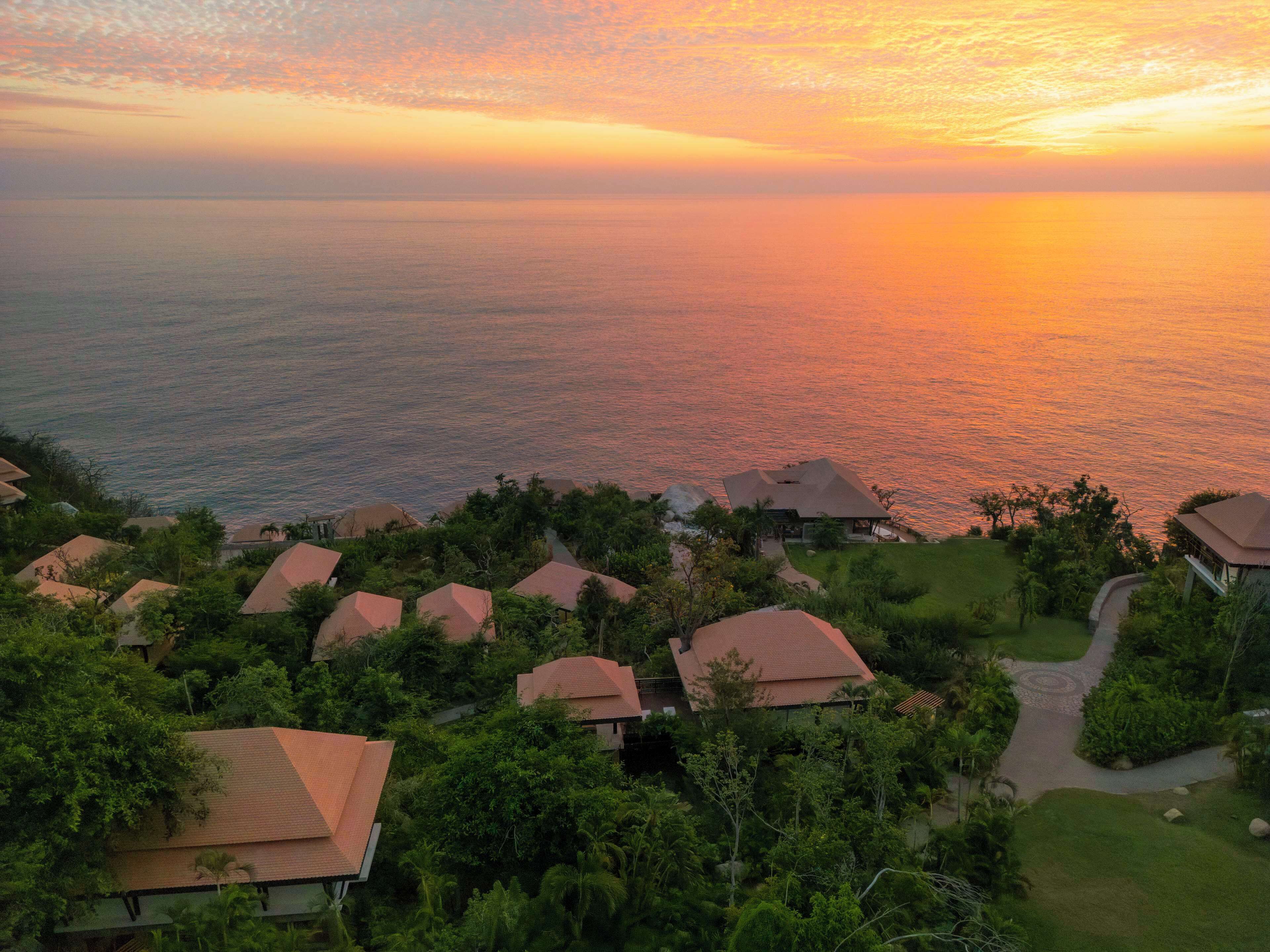 Un resort con varias edificaciones tipo bungalow, cada una con techo rojo, rodeado de exuberante vegetación y vistas a un mar tranquilo al atardecer.