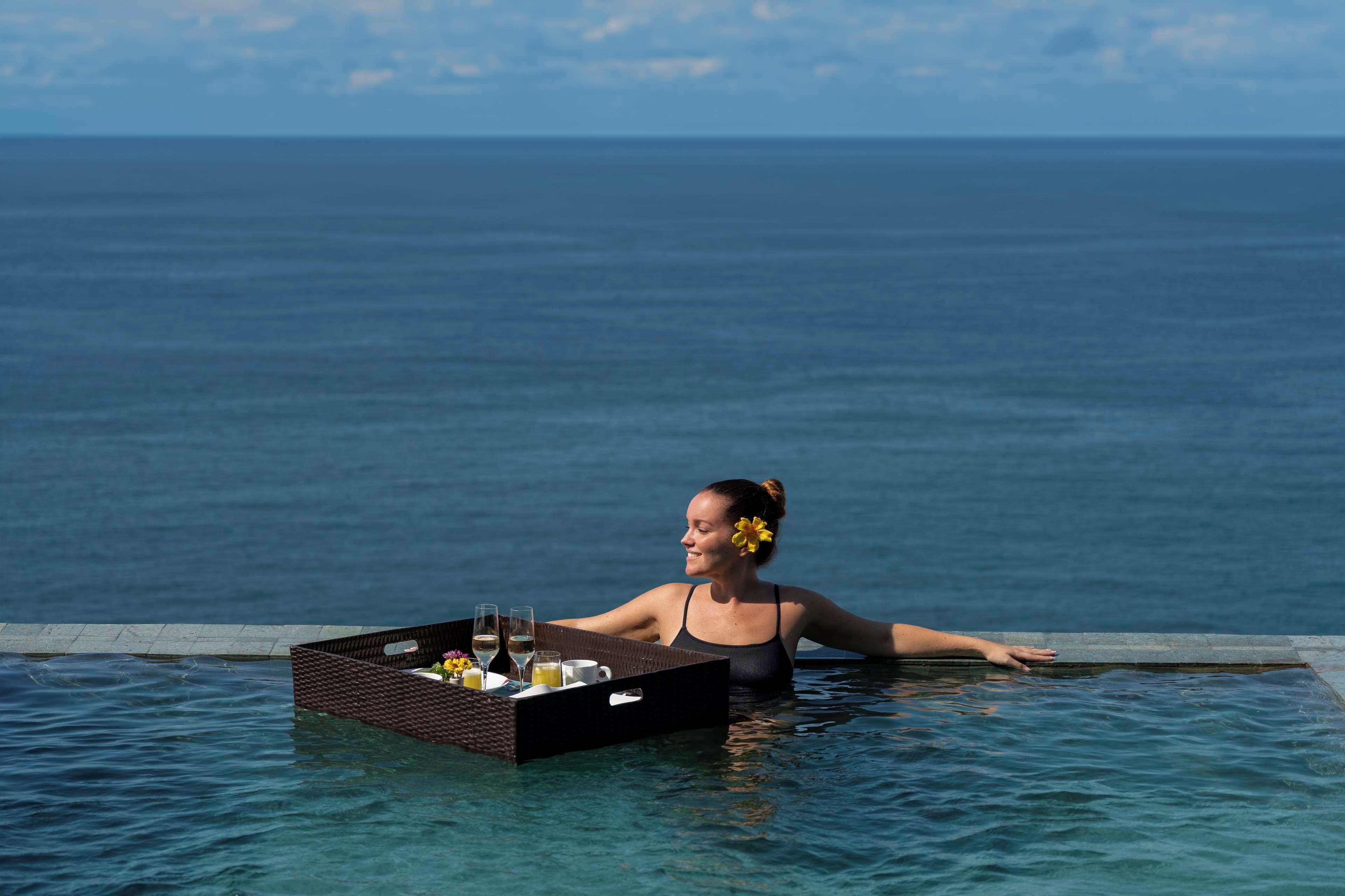 A person in a pool with a tray of drinks and a flower, overlooking the ocean.