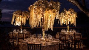 An outdoor dining area with round tables set for a meal, surrounded by chairs, and decorated with hanging floral arrangements and candles.