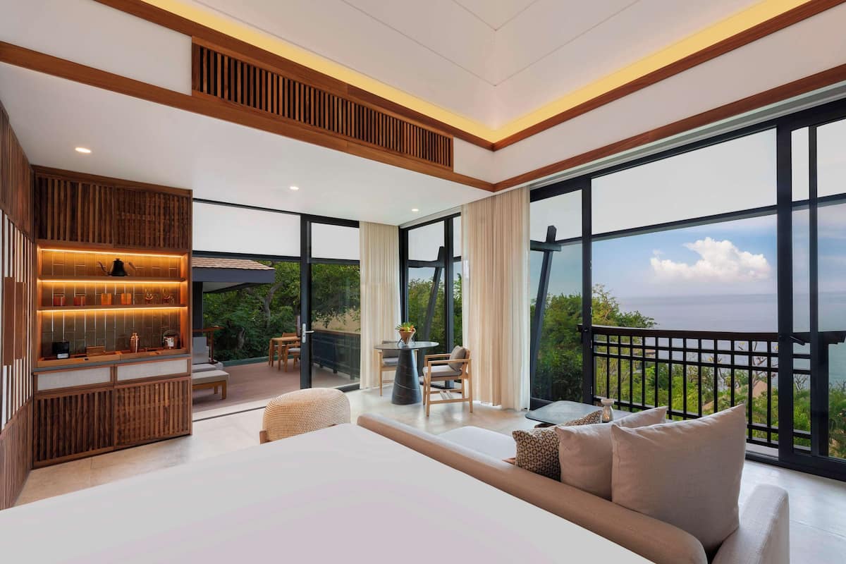 A modern bedroom with a large bed, a seating area with a table, and a view of the sea.