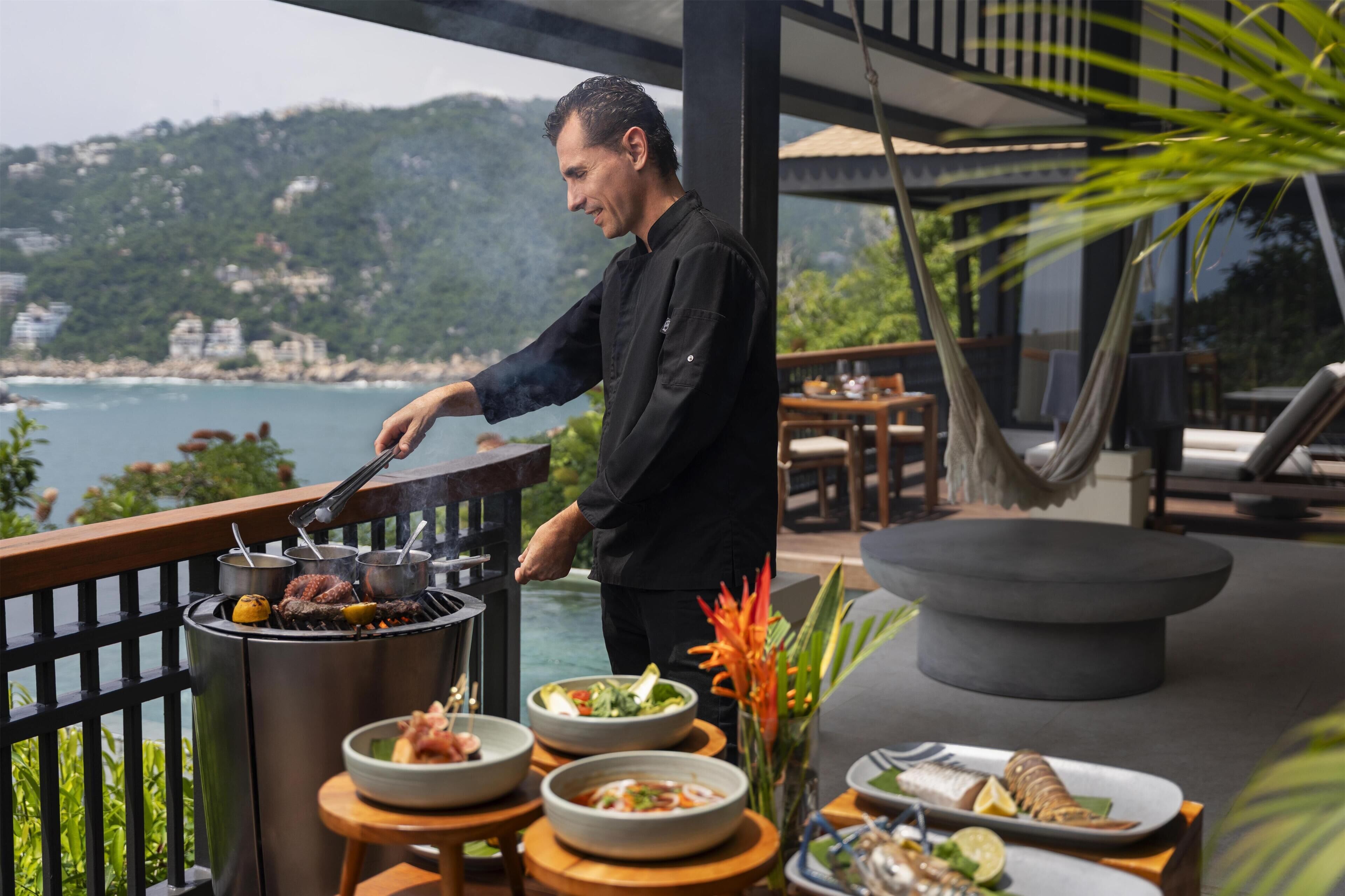 Ein Koch grillt im Freien mit Blick auf das Meer und die Hügel.