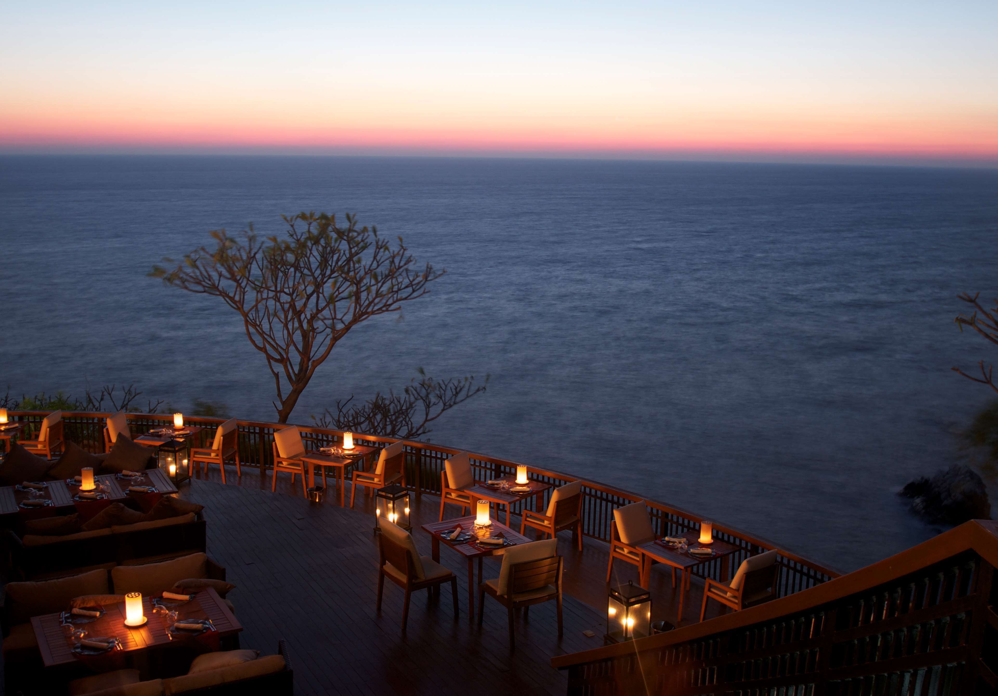 Un espacio de comedor al aire libre con mesas y sillas, con vista a una masa de agua al atardecer.
