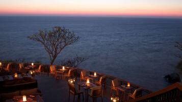 Un espacio de comedor al aire libre con mesas y sillas, con vista a una masa de agua al atardecer.