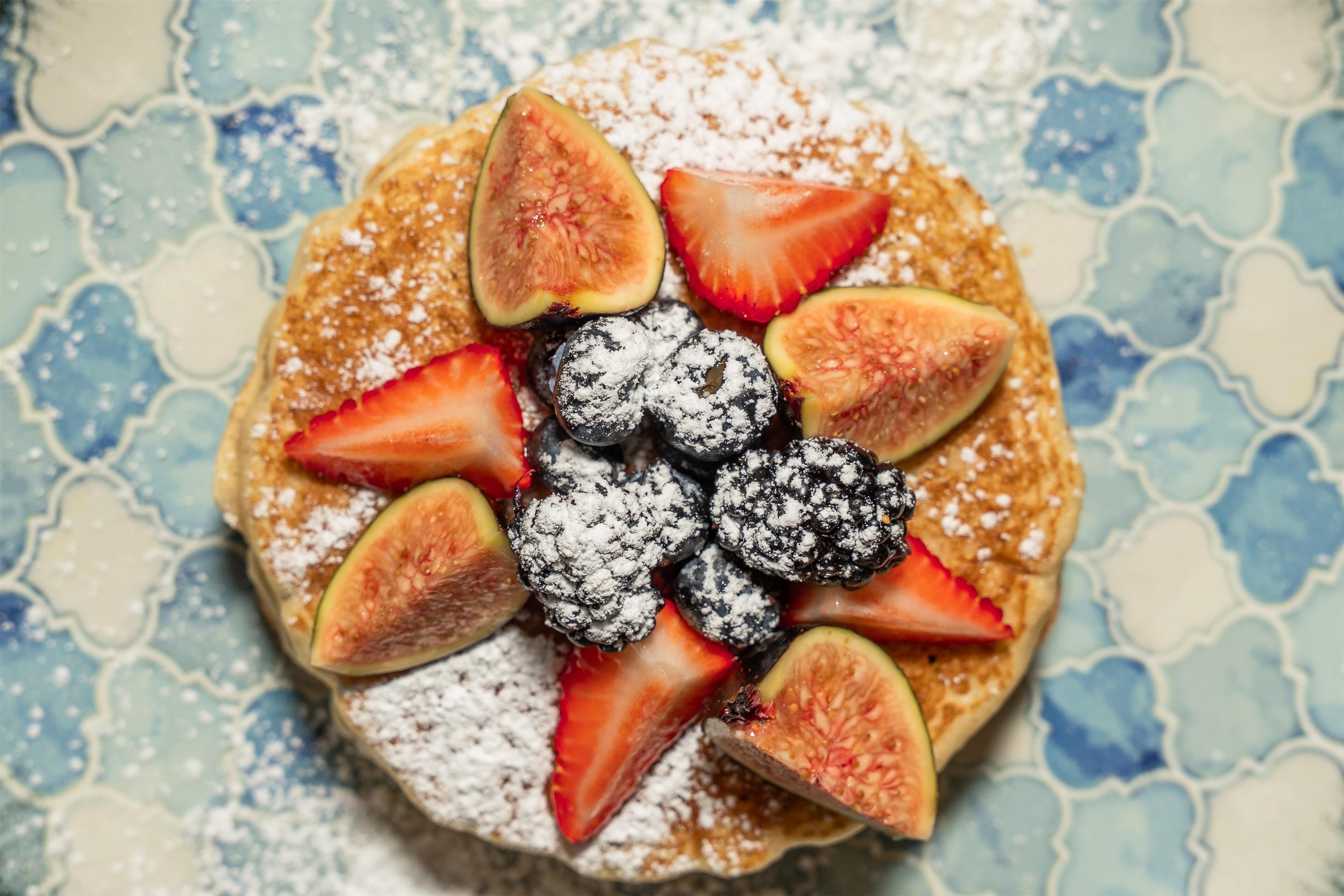 A plate of pancakes topped with strawberries, figs, blueberries, and powdered sugar.