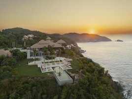 Un resort con varias construcciones, un área de piscina y vistas al océano al atardecer.