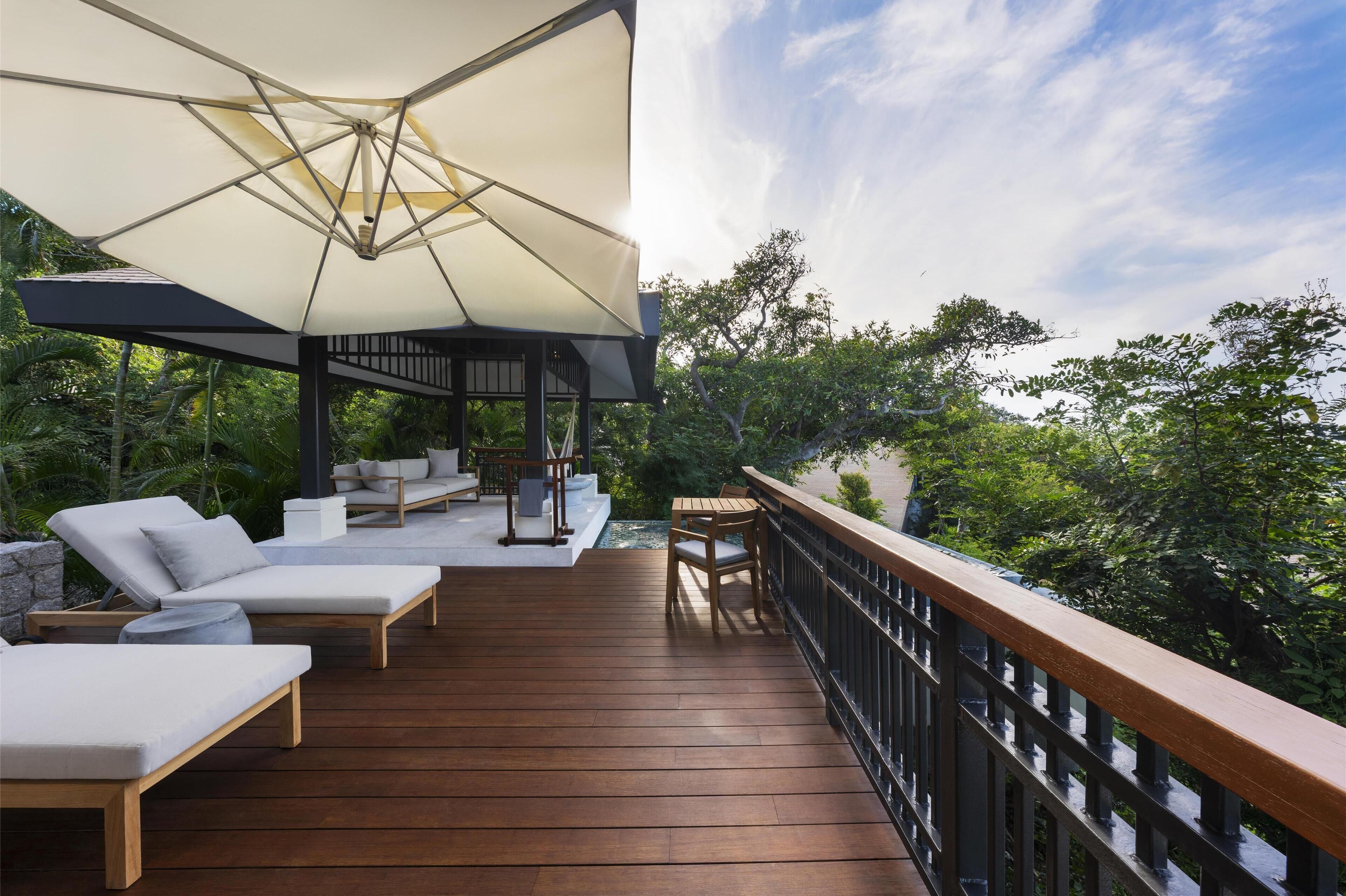 Une terrasse en bois avec des chaises longues et une table, entourée d’une rambarde et de verdure.