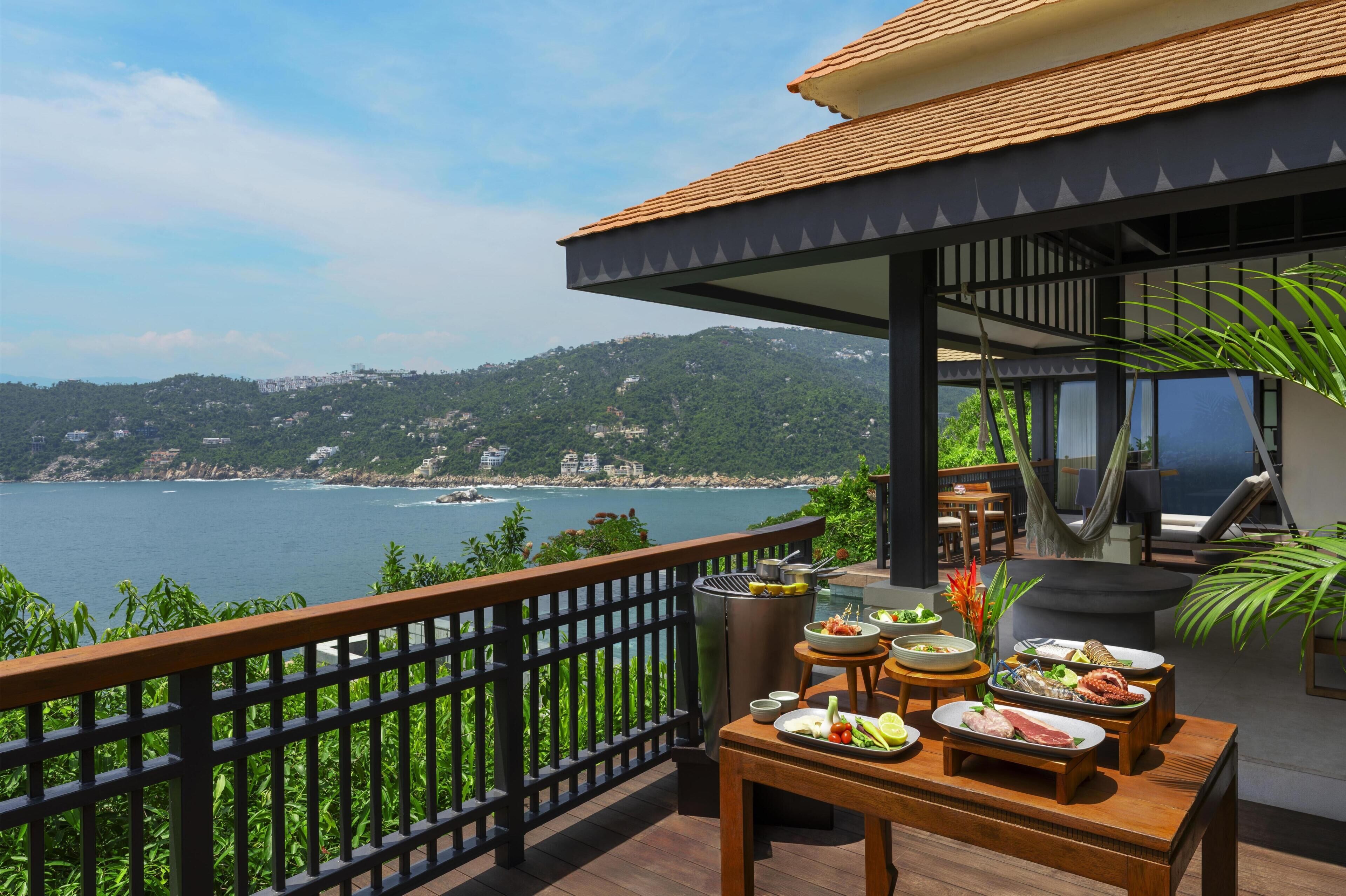 A balcony with a table set for dining, overlooking a coastal view with hills in the background.