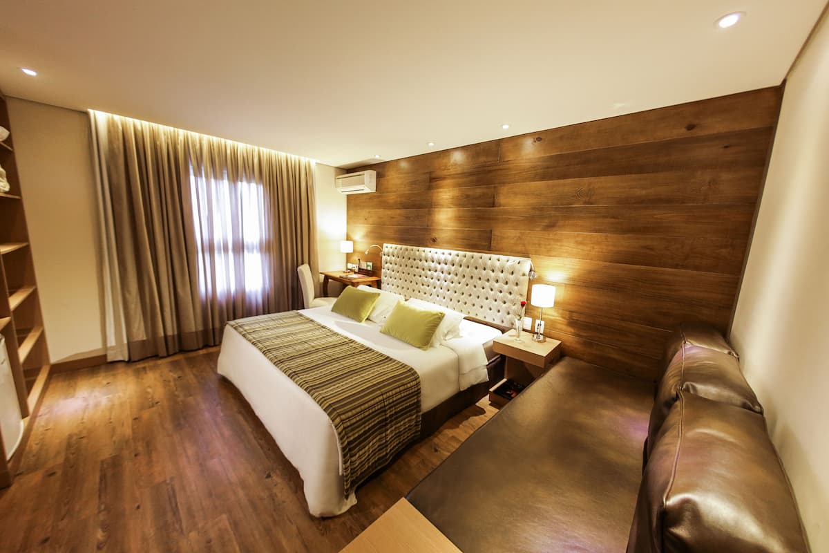 A modern hotel room with a wooden headboard, a large bed with a striped bedspread, a leather sofa, and a wooden floor.
