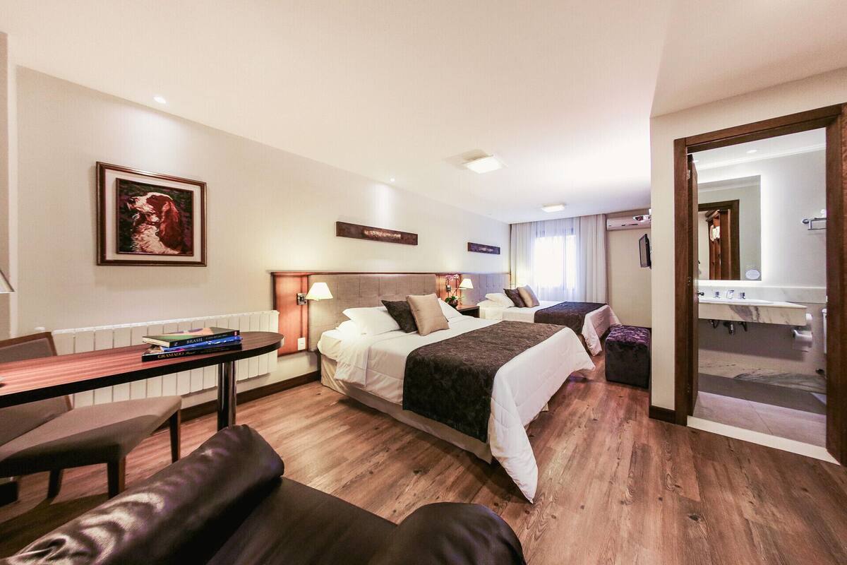A hotel room with two beds, a desk, a chair, a small table, a mirror, and a bathroom visible through an open door.