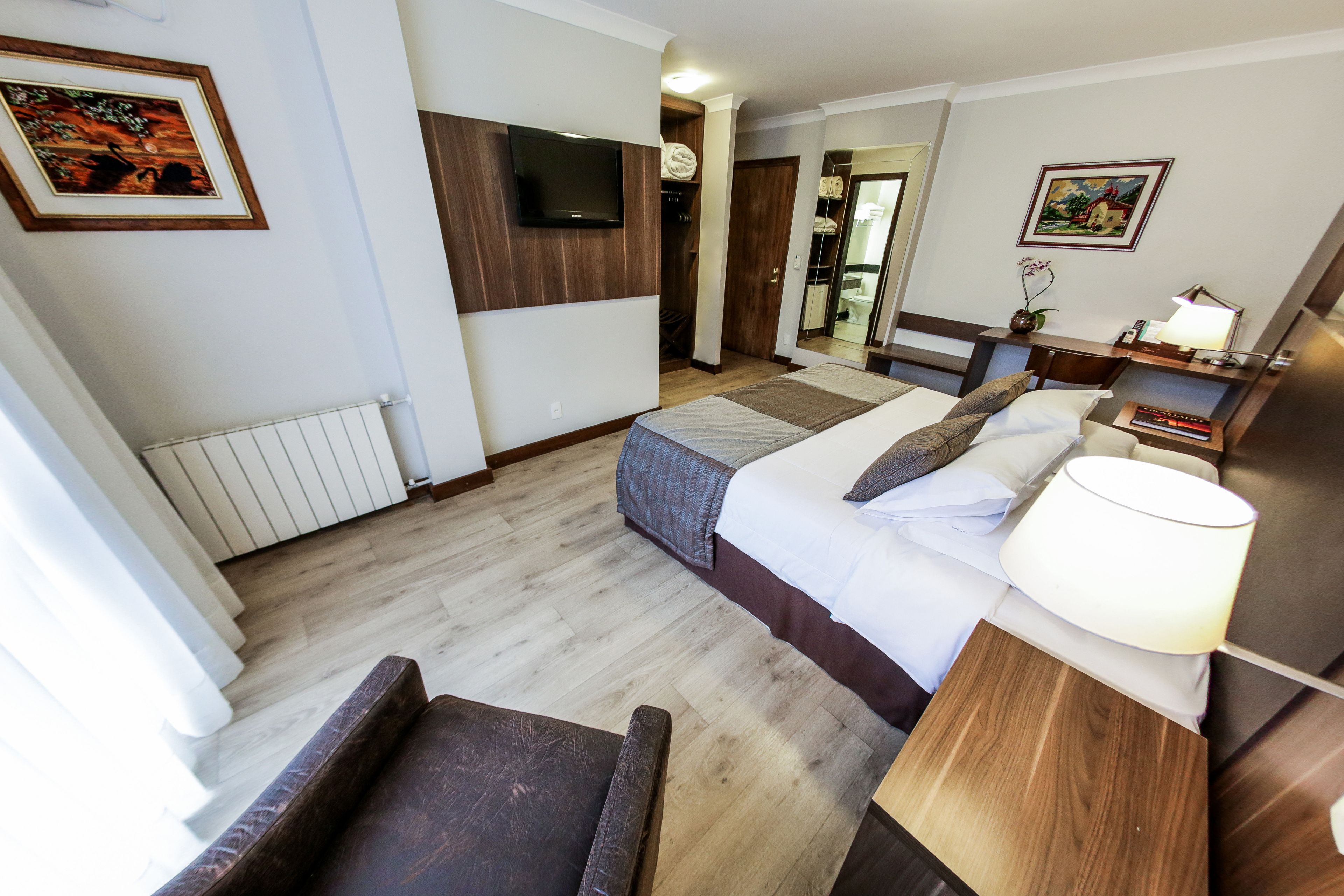 A hotel room with a bed, a chair, a bedside table with a lamp, a TV mounted on the wall, and a framed picture on the wall.