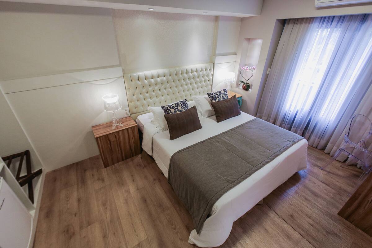 A modern hotel room with a large bed, wooden flooring, a tufted headboard, and a nightstand with a lamp.