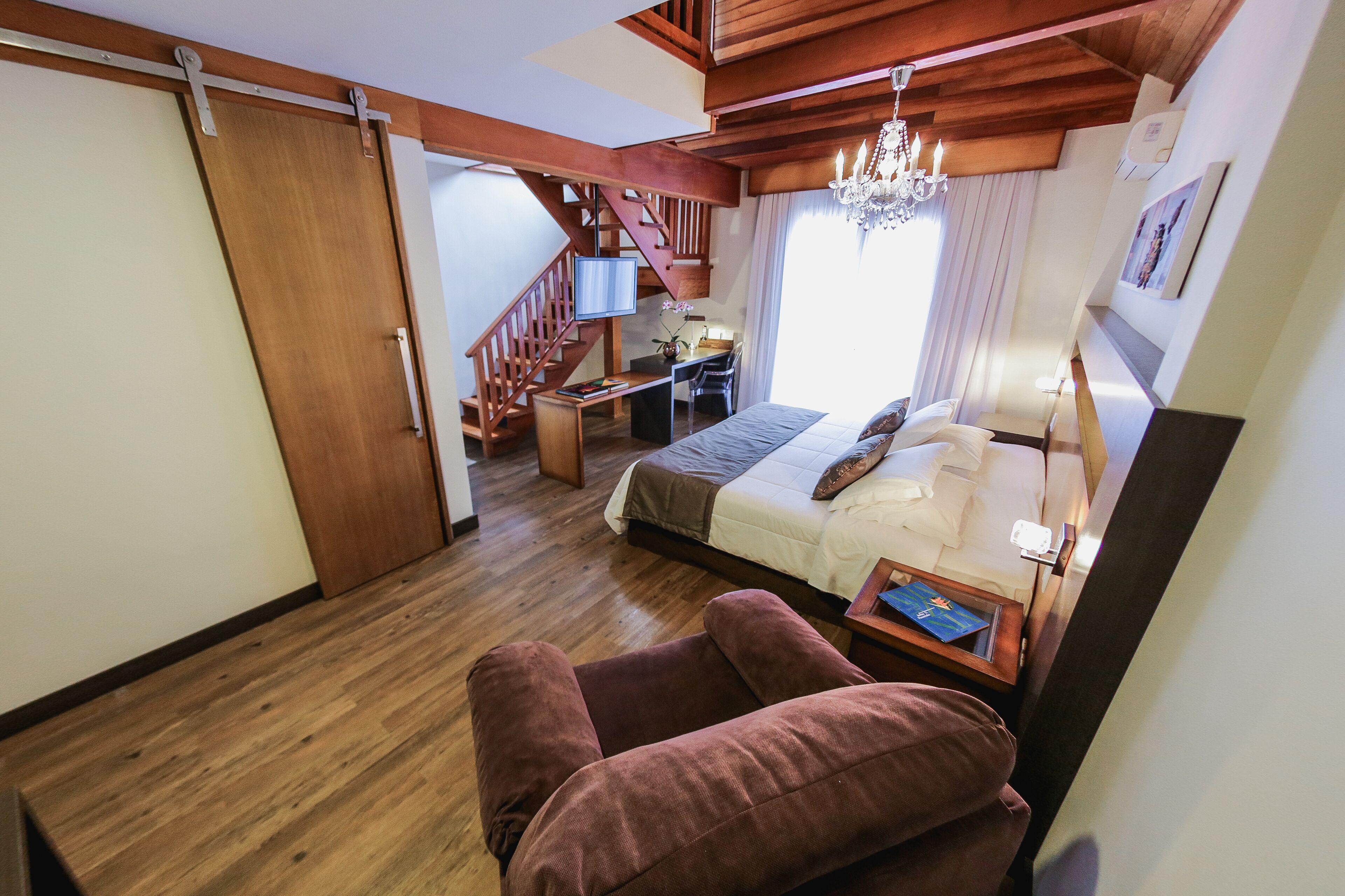 A bedroom with a bed, a desk, a chair, a chandelier, and a staircase.
