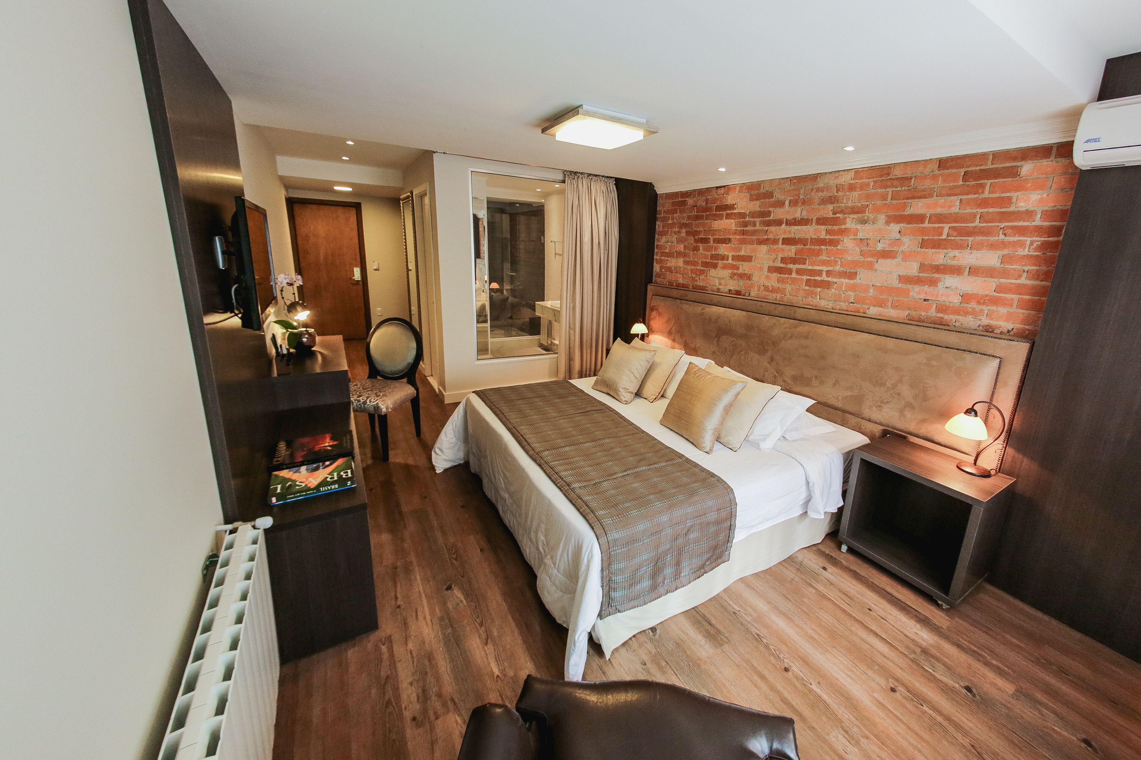 A modern hotel room with a large bed, a wooden floor, a brick accent wall, a small bedside table with a lamp, and a view of the bathroom.