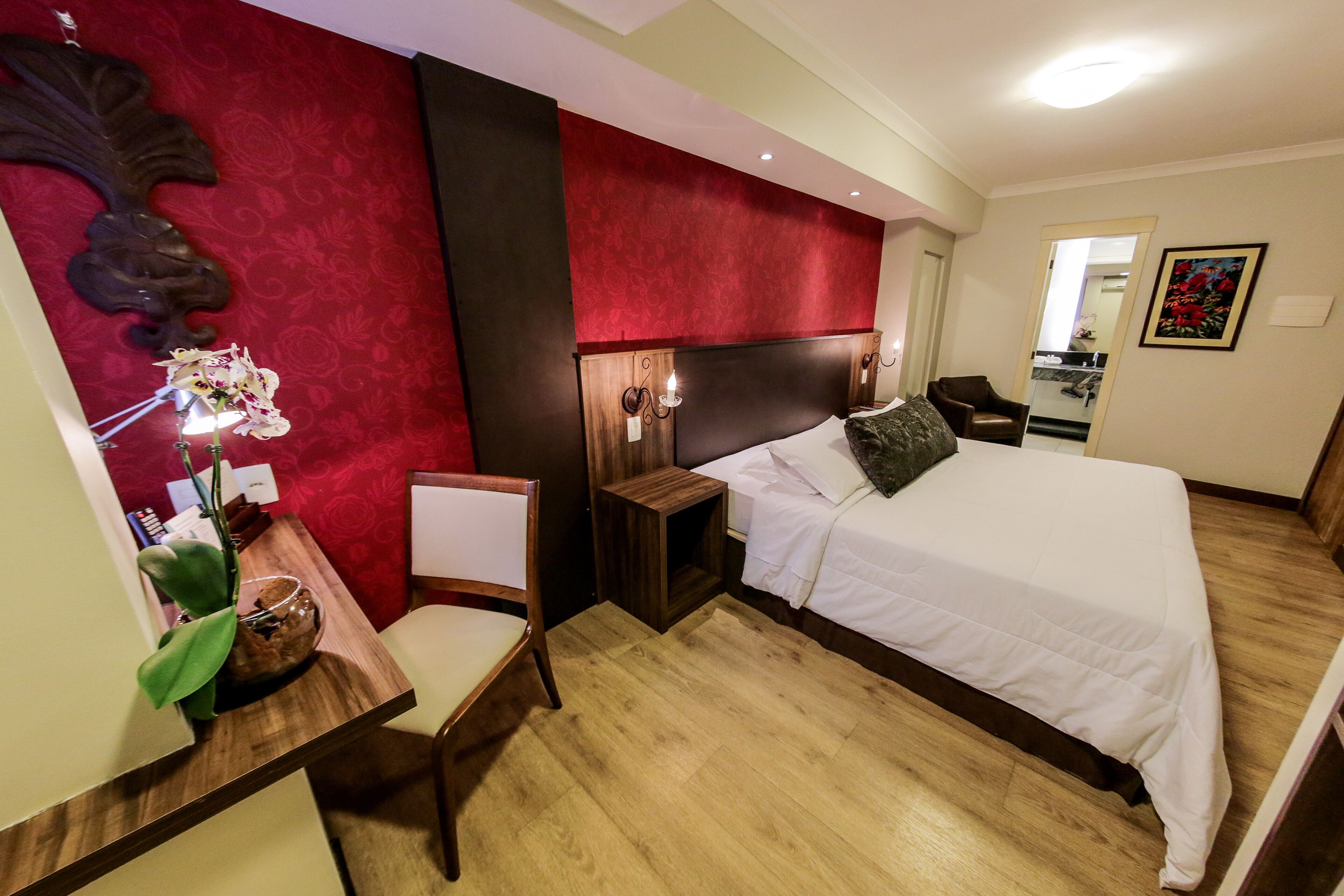 Una habitación de hotel con cama, un escritorio con silla, una mesita con un florero y un espejo en la pared.