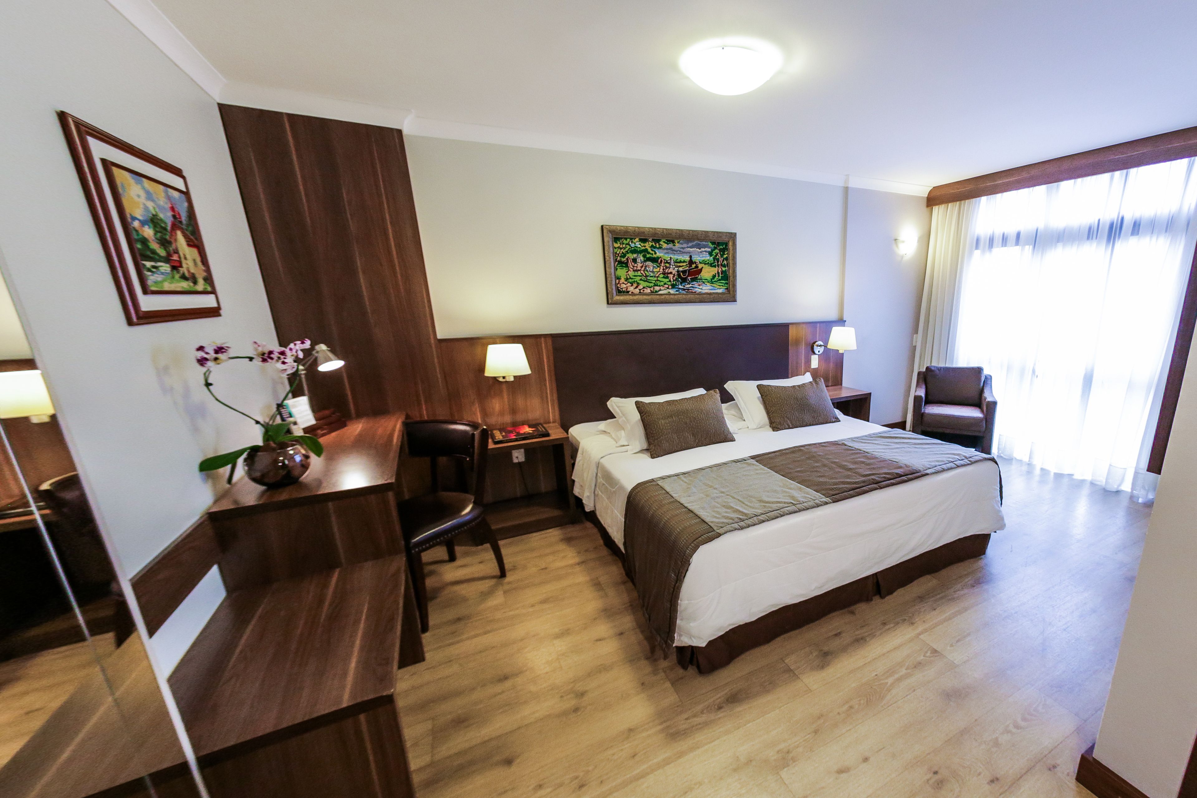 A hotel room with a large bed, a desk, a chair, and two paintings on the walls.