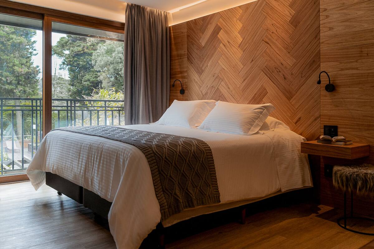 A modern bedroom with a large bed, wooden wall paneling, a small bedside table with a book, and a view of the outdoors through a sliding door.
