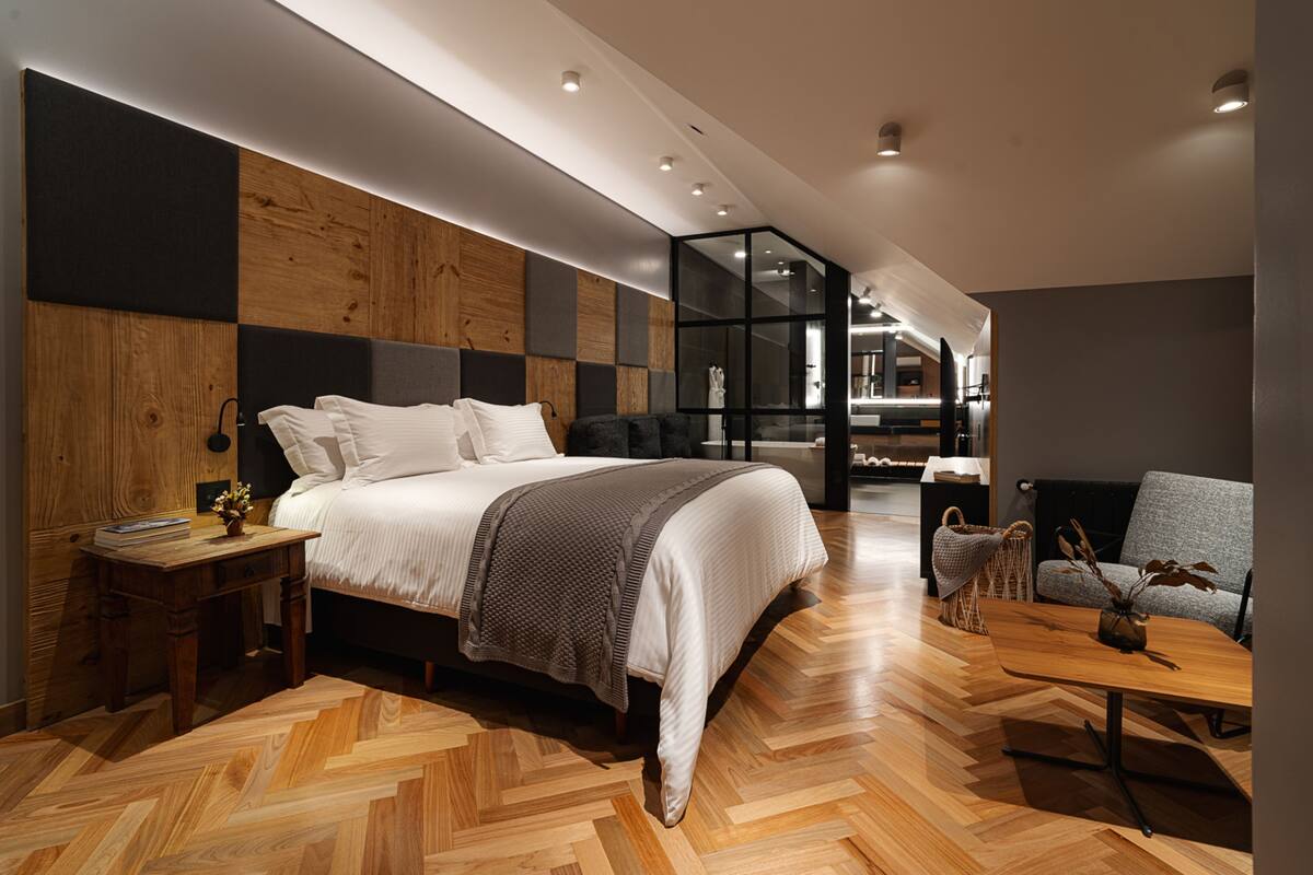 A modern bedroom with a large bed, wooden panel walls, a herringbone floor, a bedside table with a lamp, and a seating area with a sofa and a coffee table.