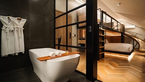 A modern bathroom with a freestanding bathtub, wooden flooring, and a view of a wooden staircase leading to a loft bedroom. - Bavaria Sport Hotel (Gramado)