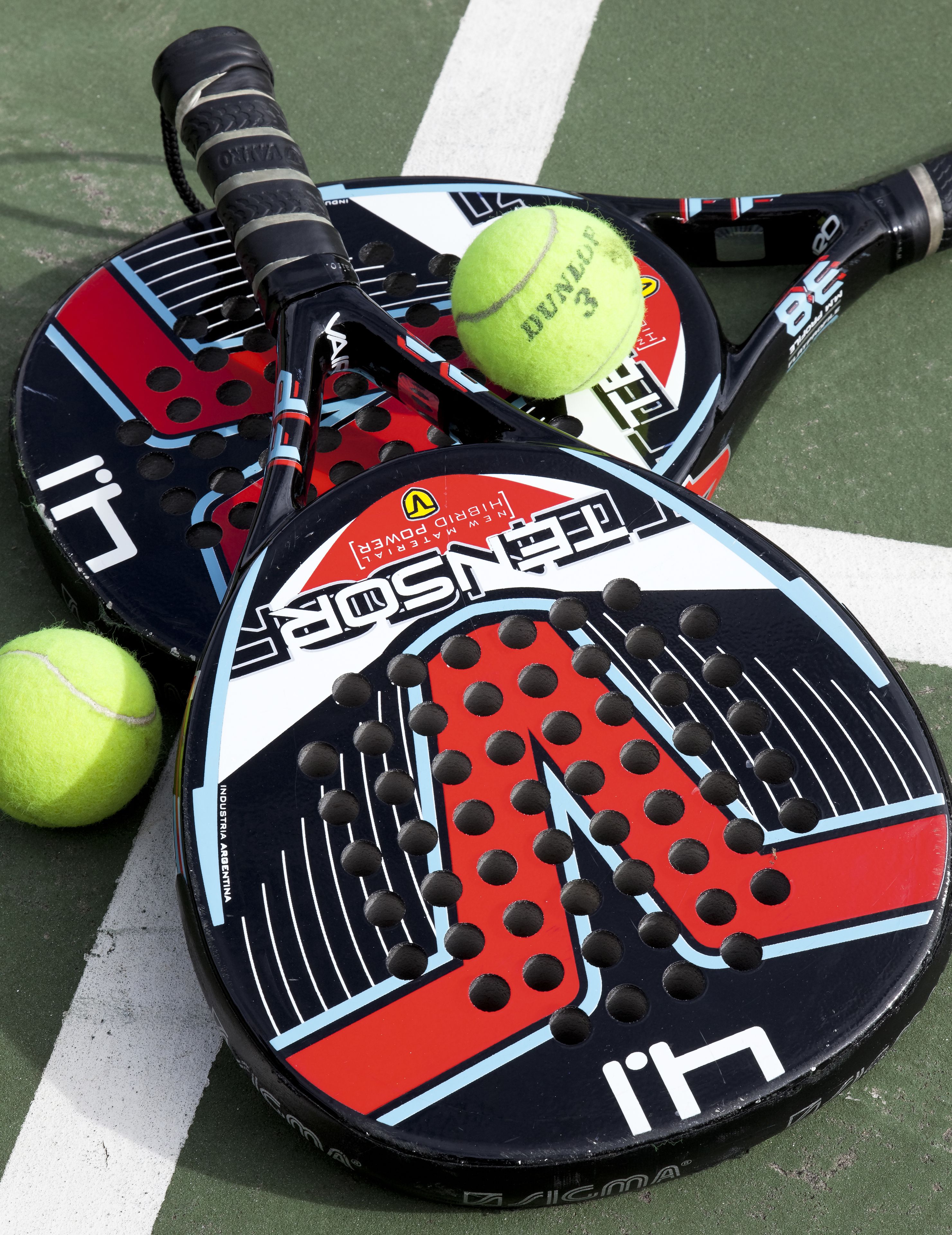 Dos raquetas de tenis con un diseño rojo, negro y azul, y dos pelotas de tenis sobre una cancha verde.