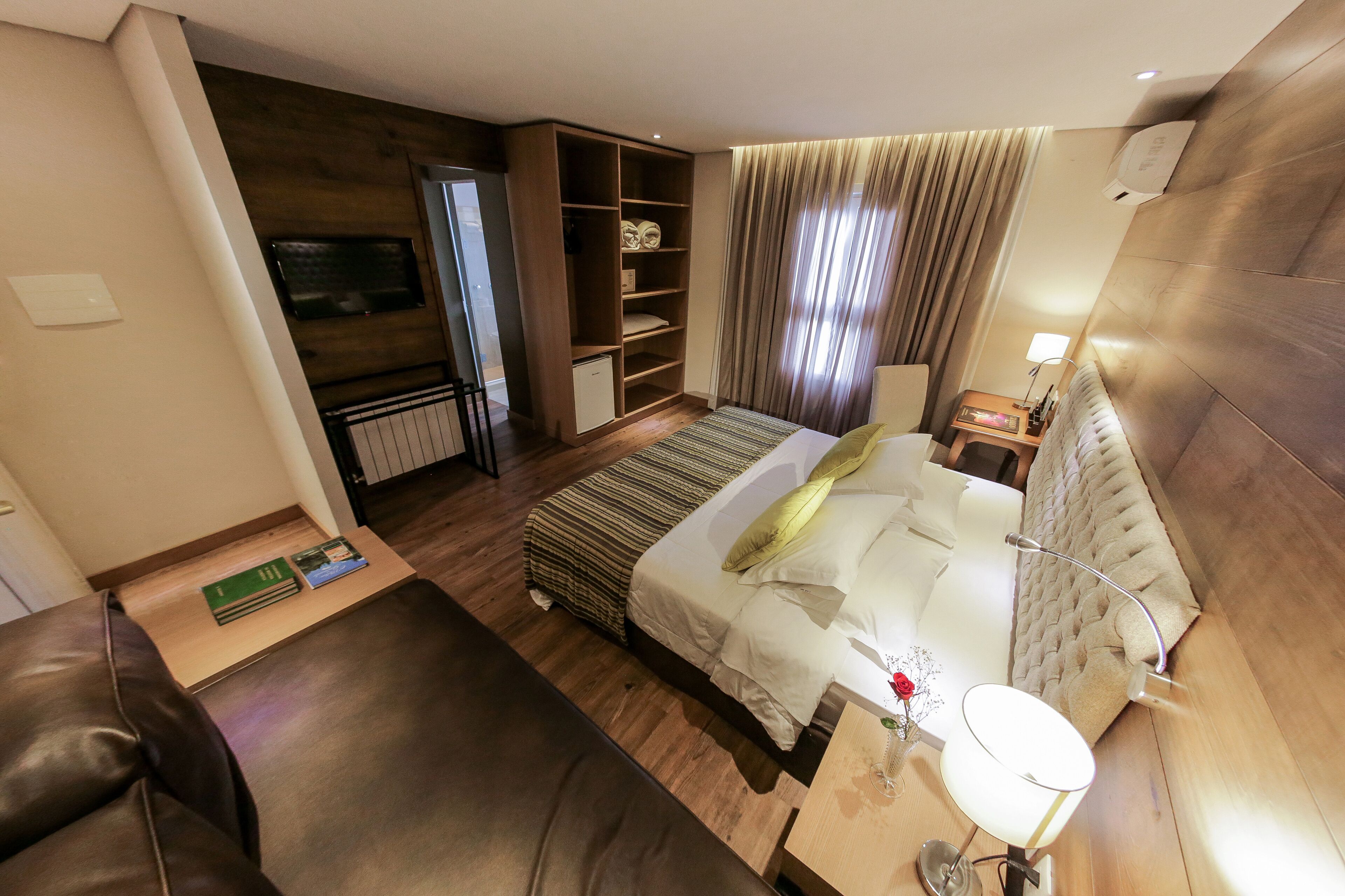 A modern hotel room with a bed, a seating area, a TV, a wardrobe, and a small kitchenette.