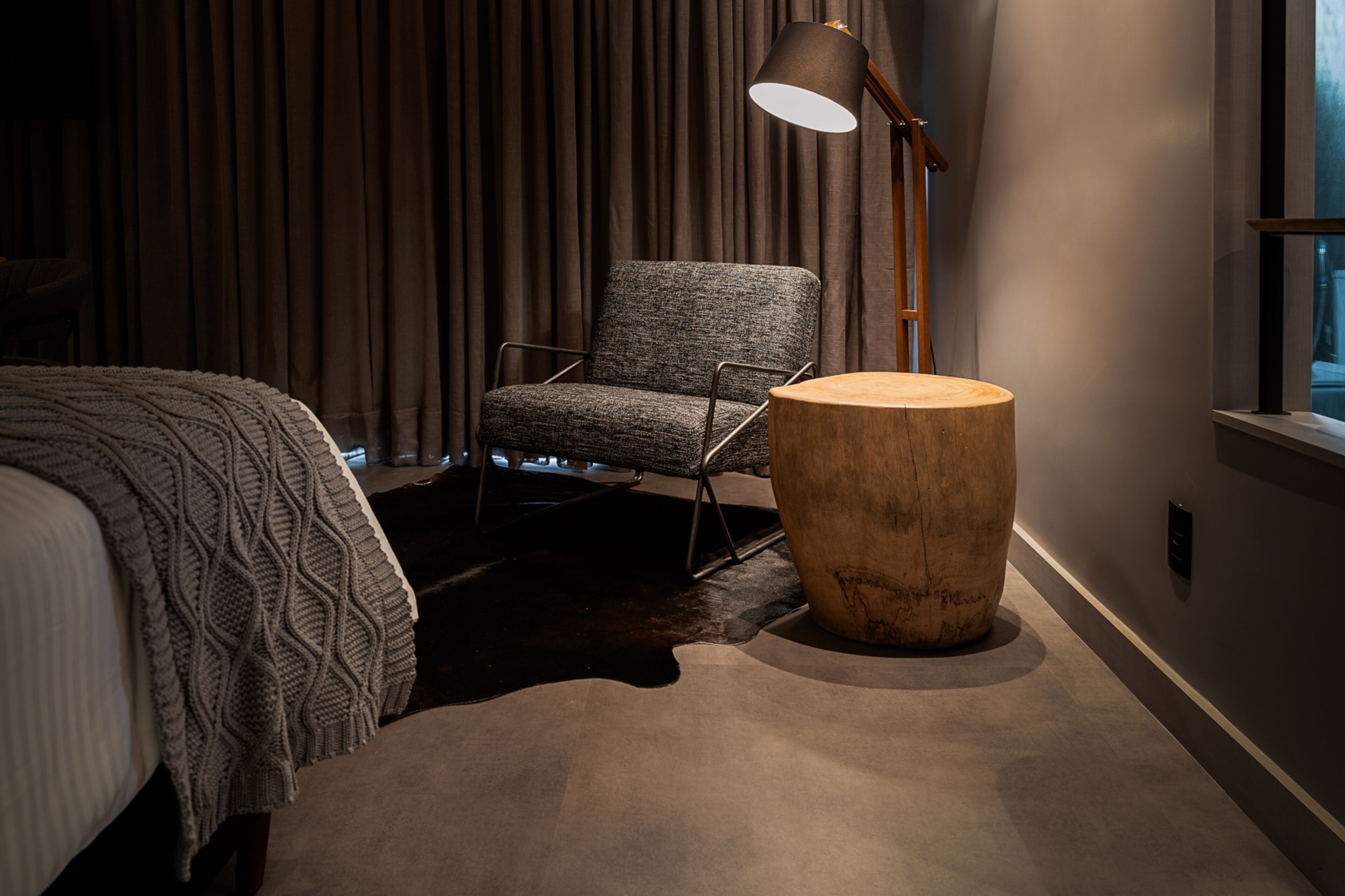 A bedroom with a bed, a chair, a wooden side table, and a floor lamp.