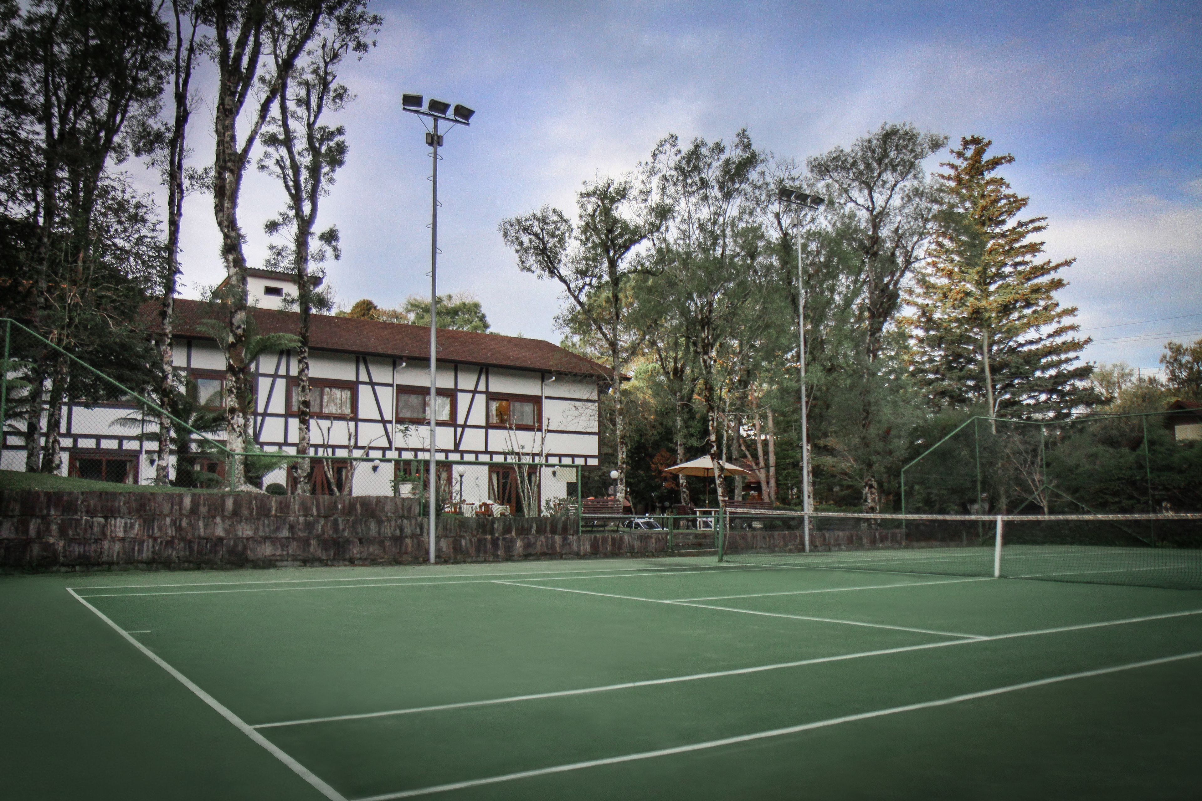 Un court de tennis en plein air, doté d’un filet, entouré d’arbres et bordé par un bâtiment au toit rouge.
