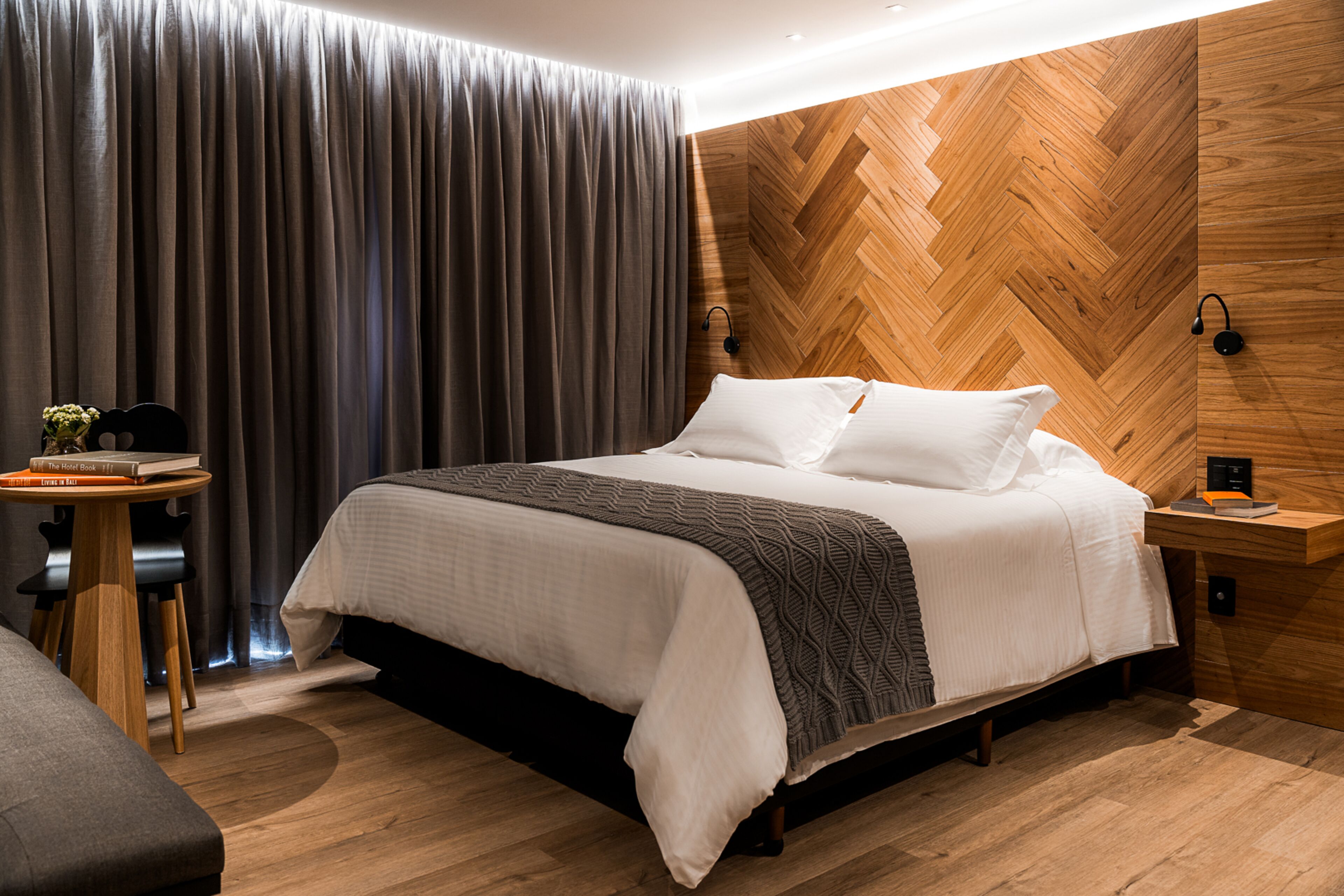 A modern hotel room with a large bed, a wooden herringbone wall, a small table with a chair, and a curtain.