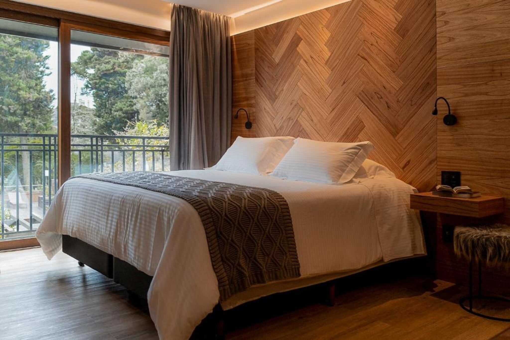 A bedroom with a large bed, a wooden herringbone wall, a nightstand with a lamp, and a view of trees through a sliding door.