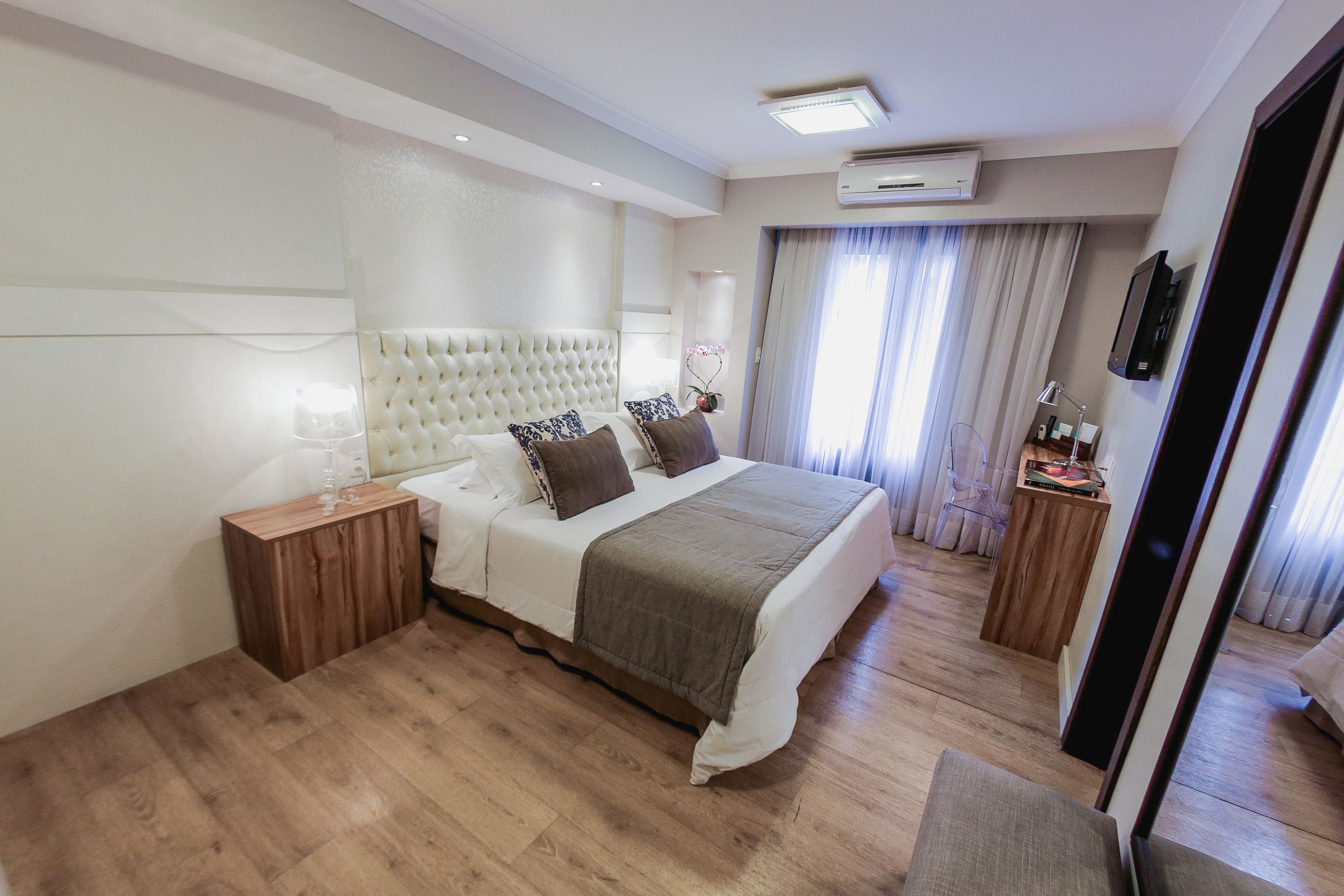 Quarto de hotel moderno com uma cama grande, piso de madeira, painel de cabeceira embutido, mesa de cabeceira com abajur e TV na parede.