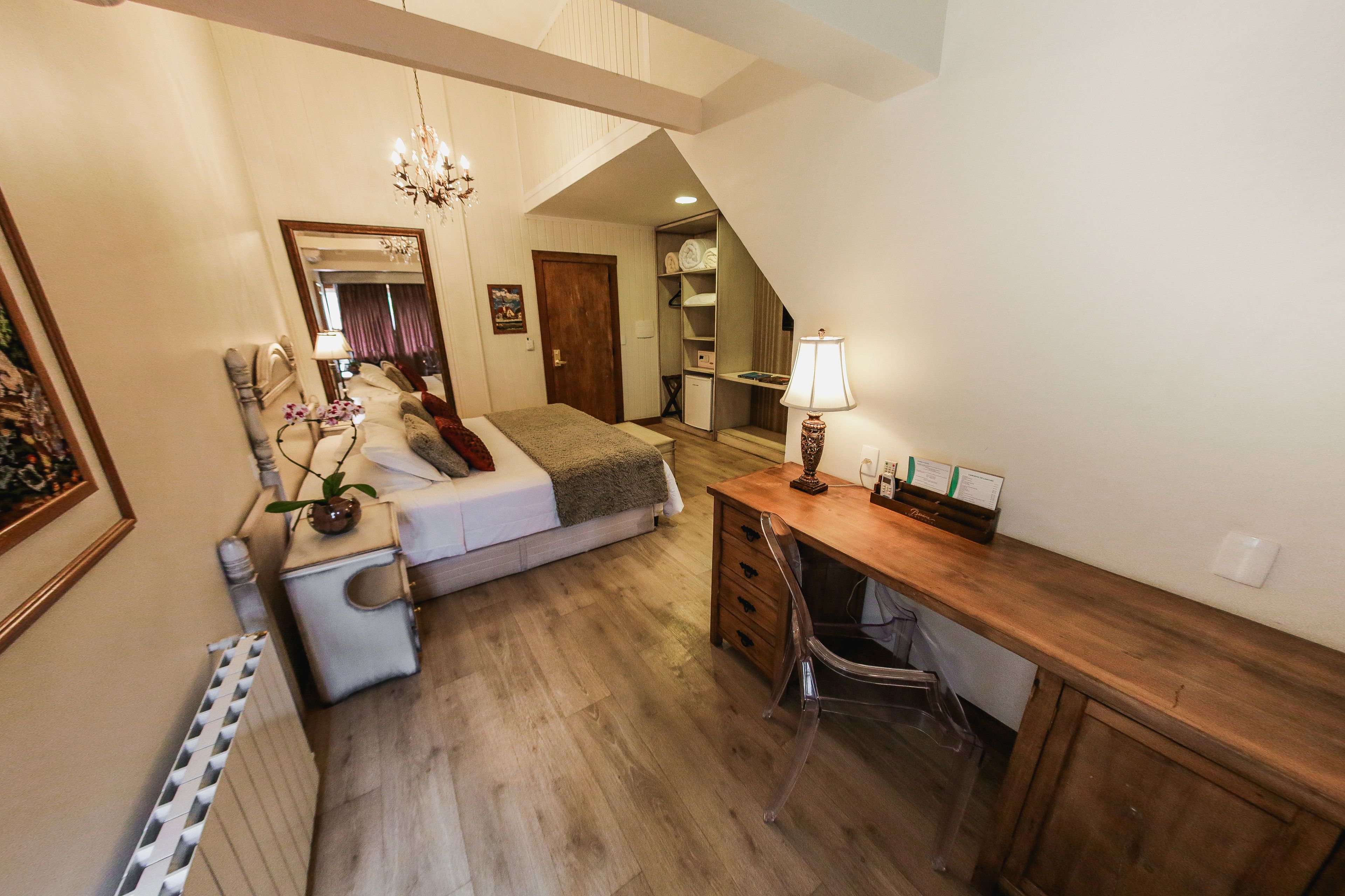 A bedroom with a bed, bedside table, lamp, and a desk with a chair. There is a mirror on the wall and a radiator on the floor.