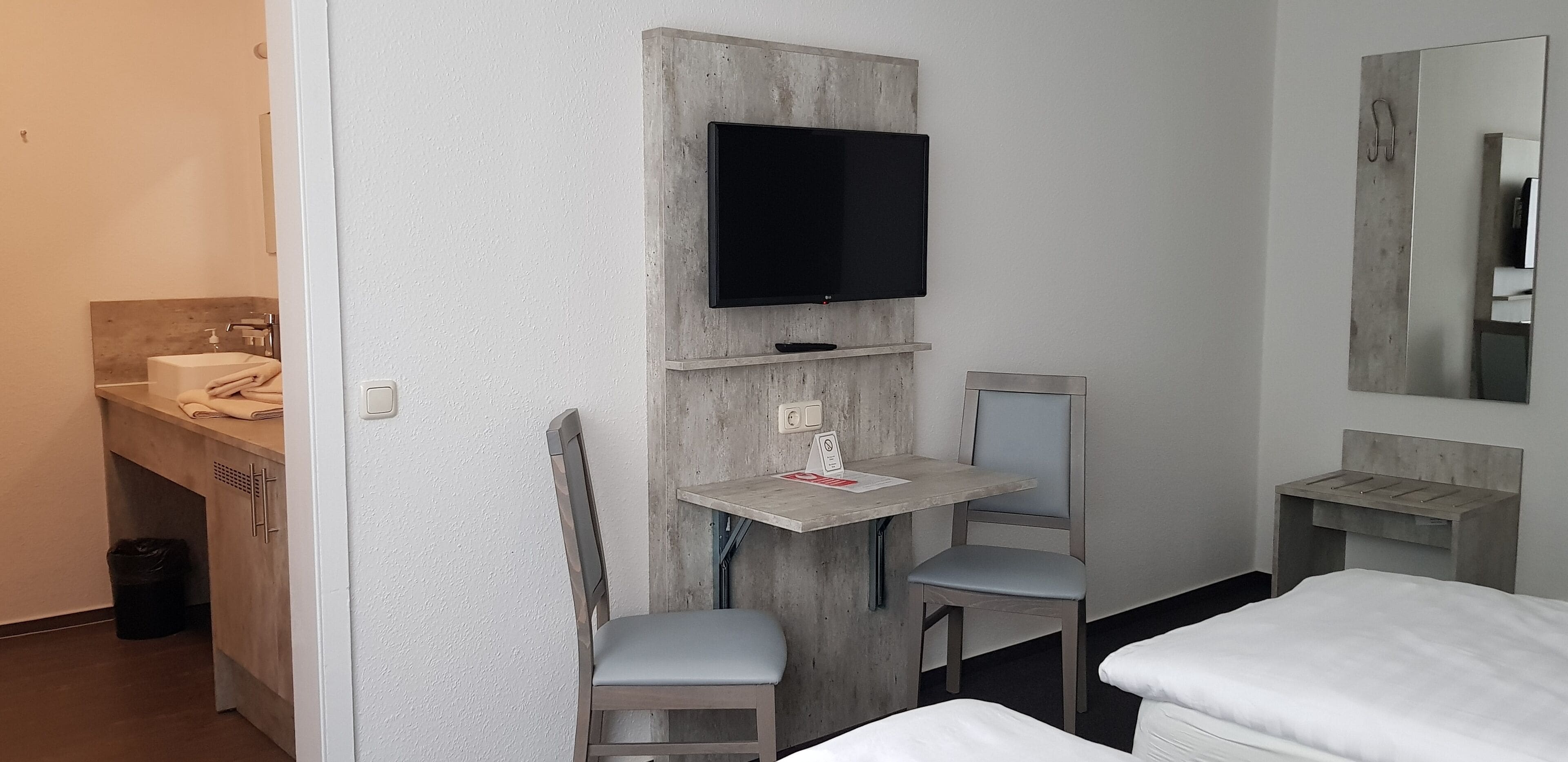Una habitación de hotel compacta con una cama, un escritorio con silla, un televisor montado en la pared, una mesita y un espejo.